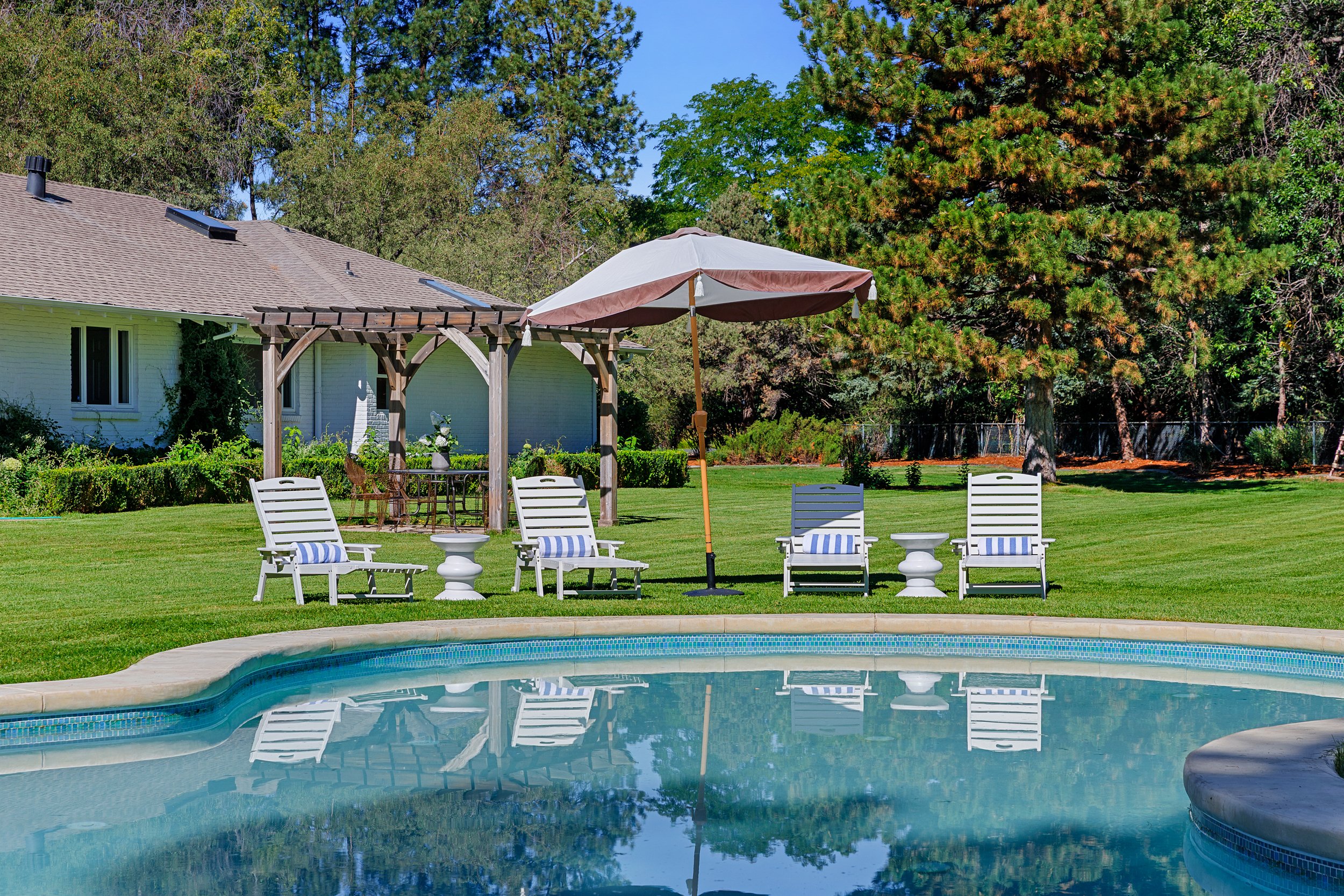 An outdoor scene featuring a swimming pool with clear blue water, surrounded by a green lawn. There are multiple white lounge chairs with blue-striped towels, a large patio umbrella, and a wooden pergola. In the background, there is a white house wit