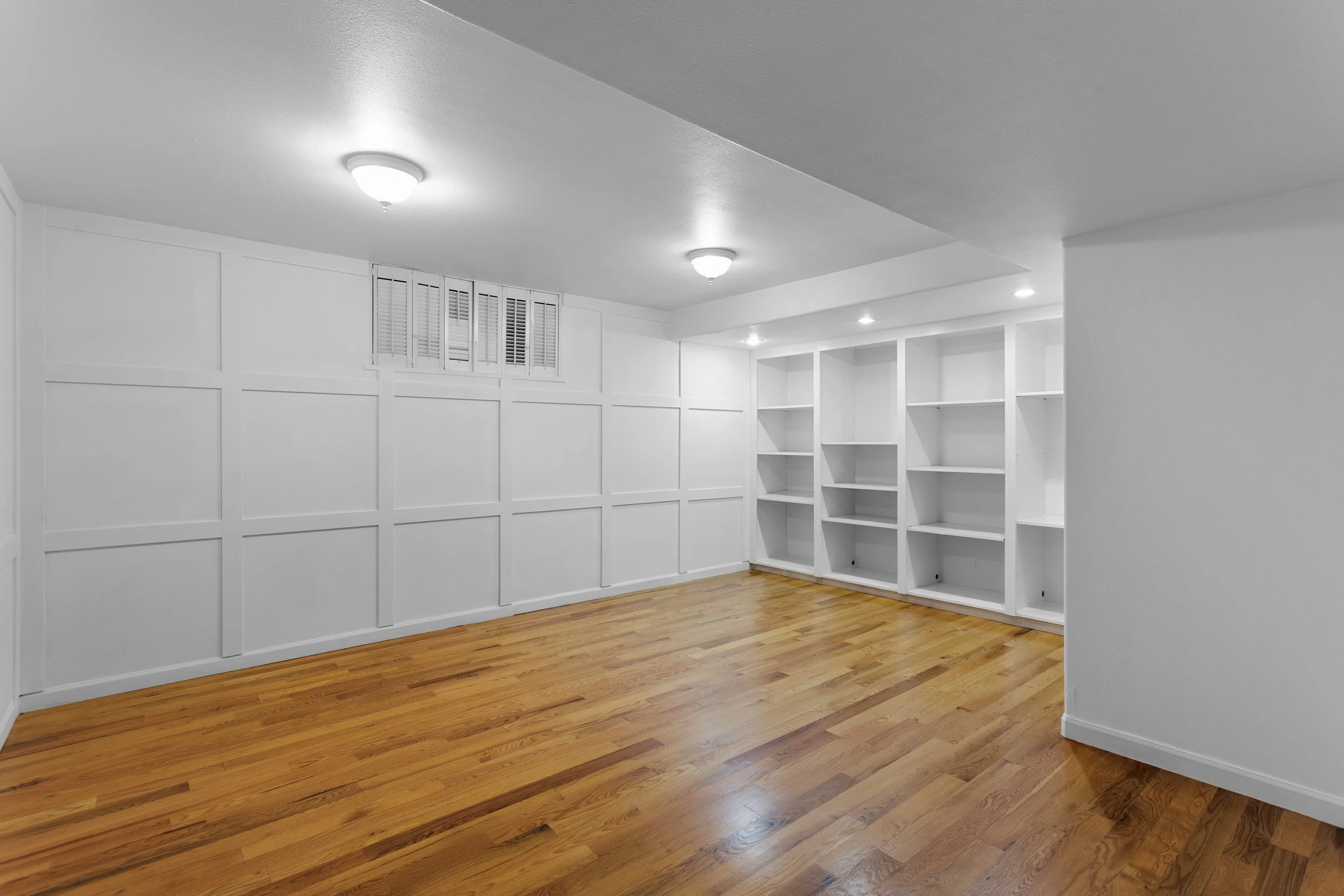 Empty basement room with white walls, built-in white bookshelves, wood laminate flooring, and ceiling lights.