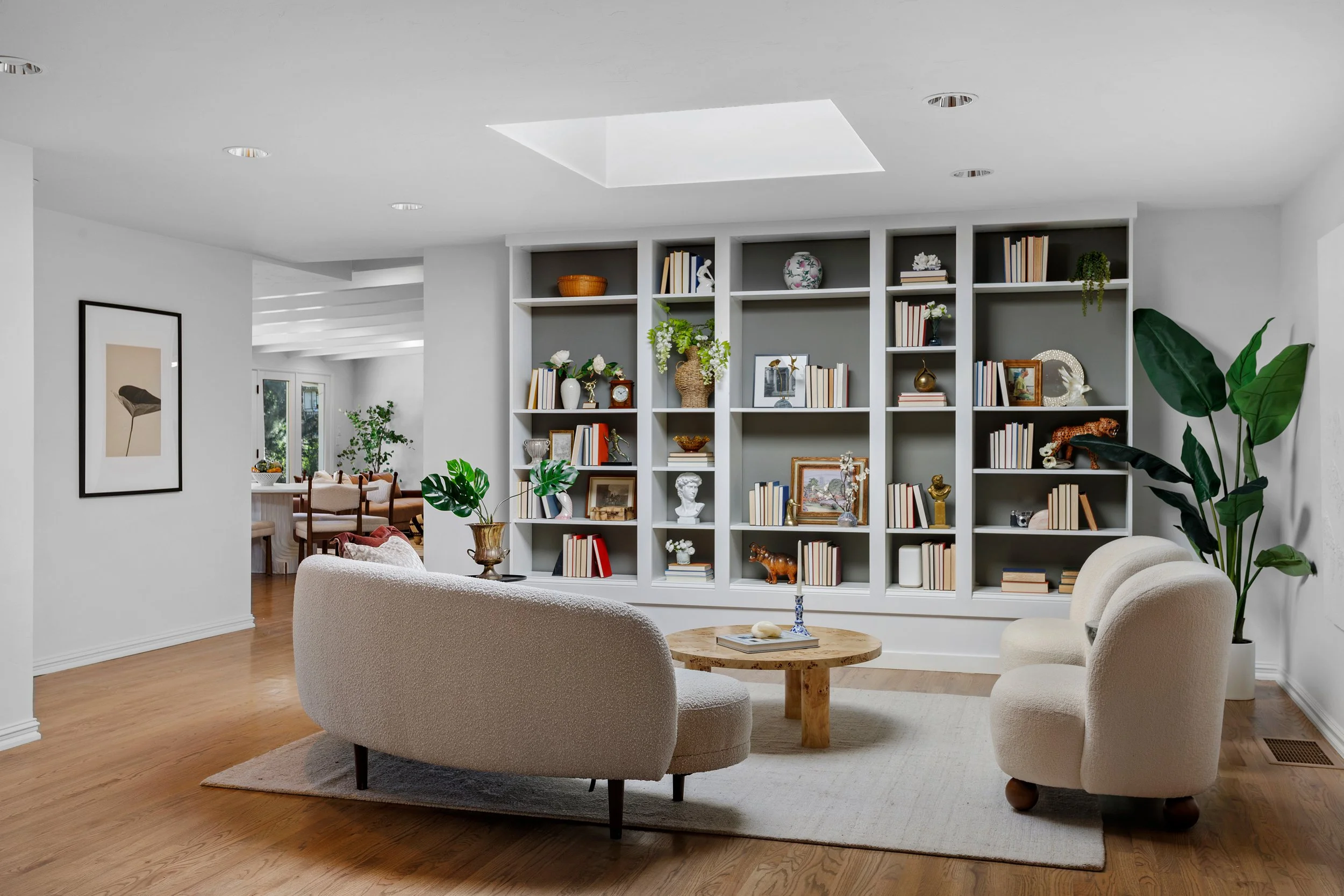 Living room with white walls, a built-in bookshelf filled with books and decorative items, a round coffee table, and two beige armchairs. There are large green plants and framed artwork on the walls. A dining area is visible in the background.