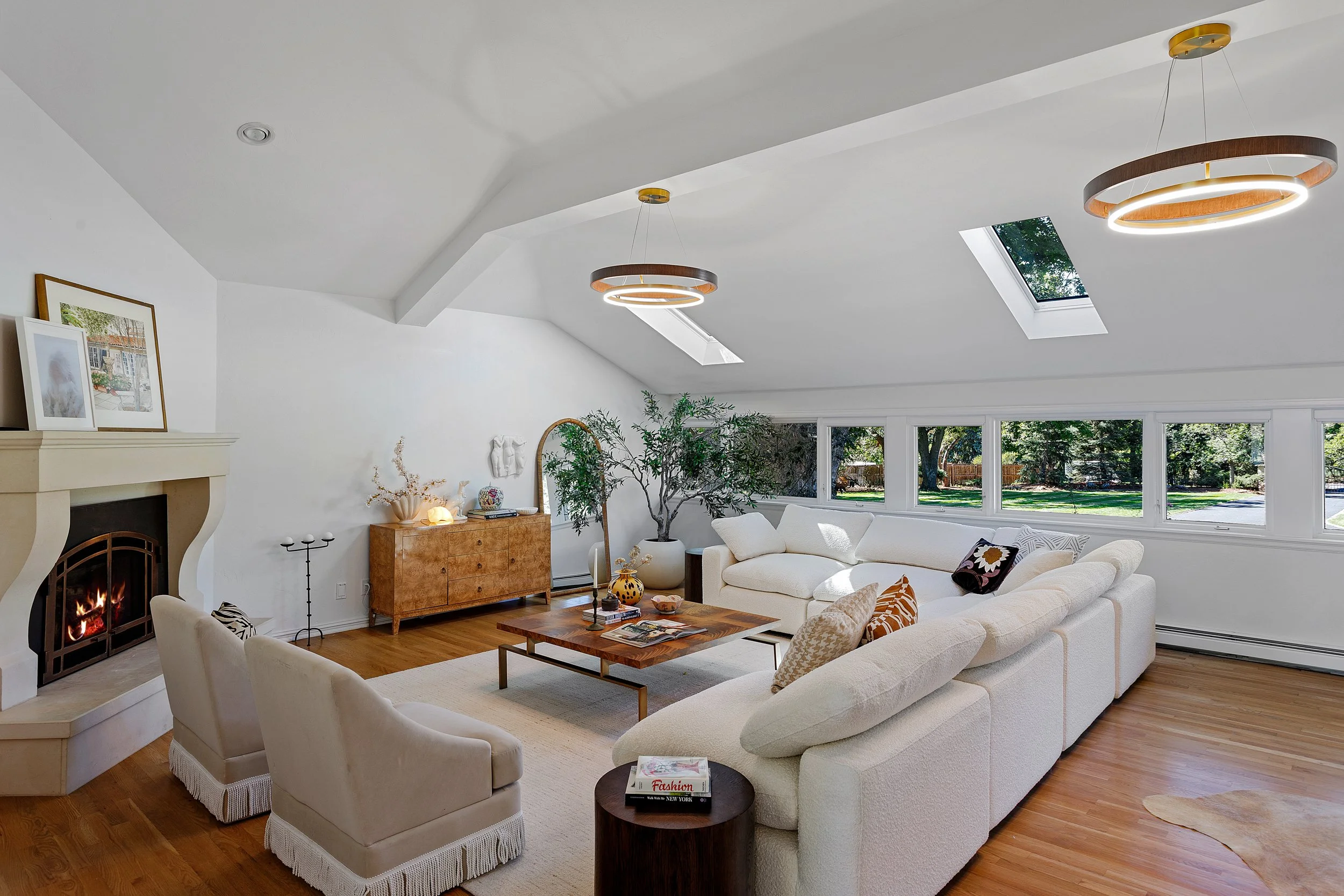A spacious, bright living room with white walls and a vaulted ceiling, featuring two skylights and modern circular hanging light fixtures. There is a fireplace on the left, a beige sectional sofa with throw pillows, a wooden coffee table with magazin