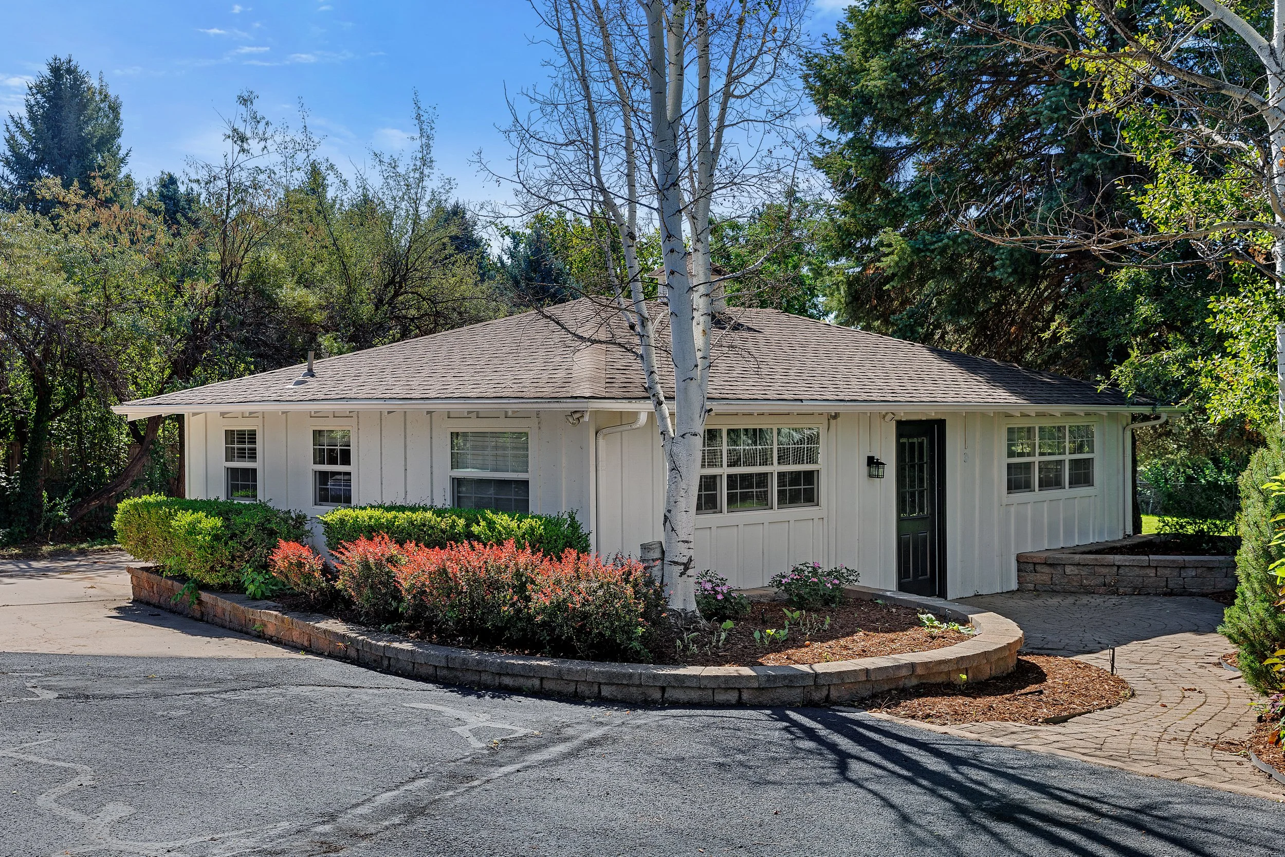 A small white house with a brown roof, surrounded by trees and bushes, with a paved driveway and flower bed in the front yard.