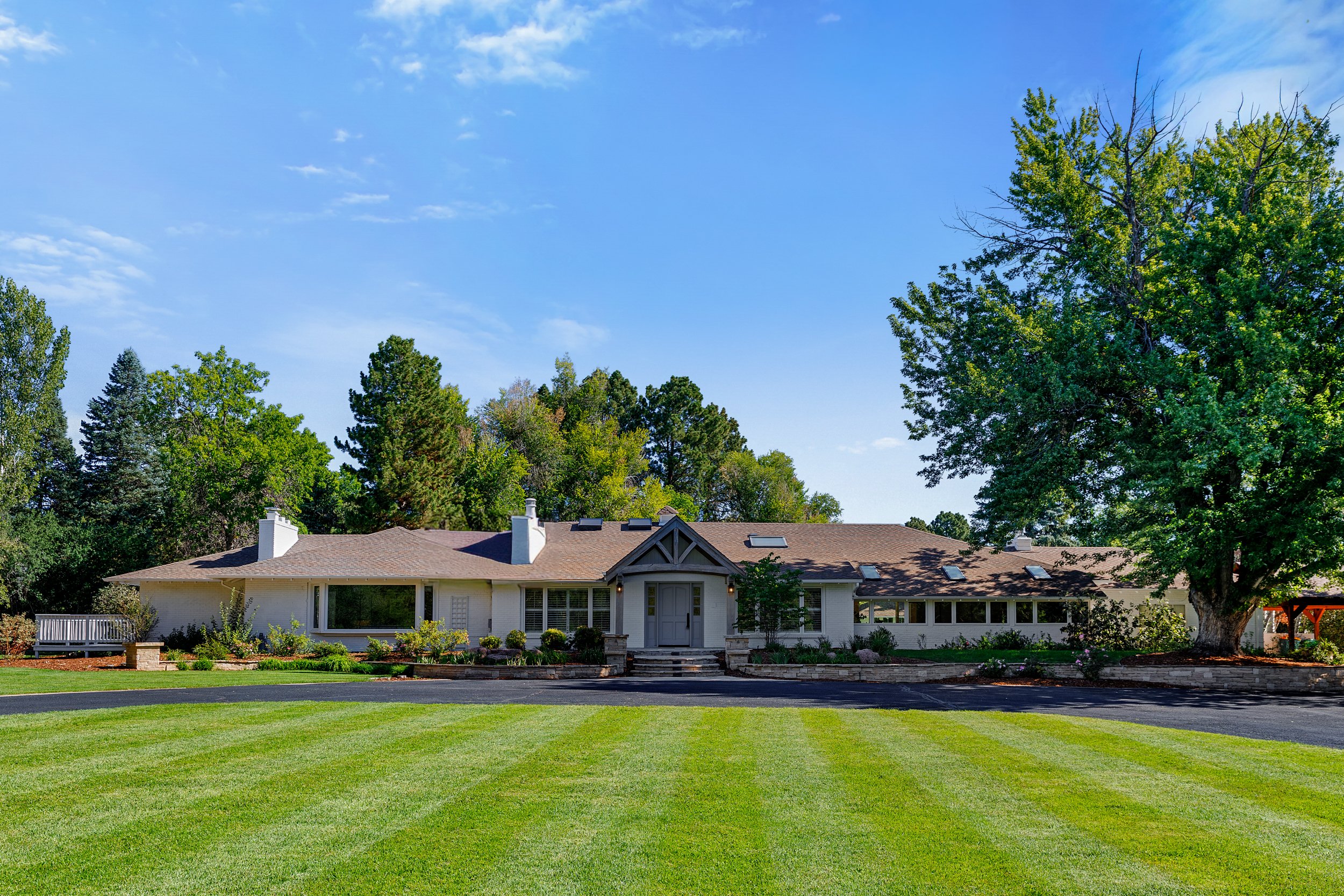 Rear view of a white house with a brown roof, surrounded by a well-maintained lawn and tall green trees under a partly cloudy blue sky.