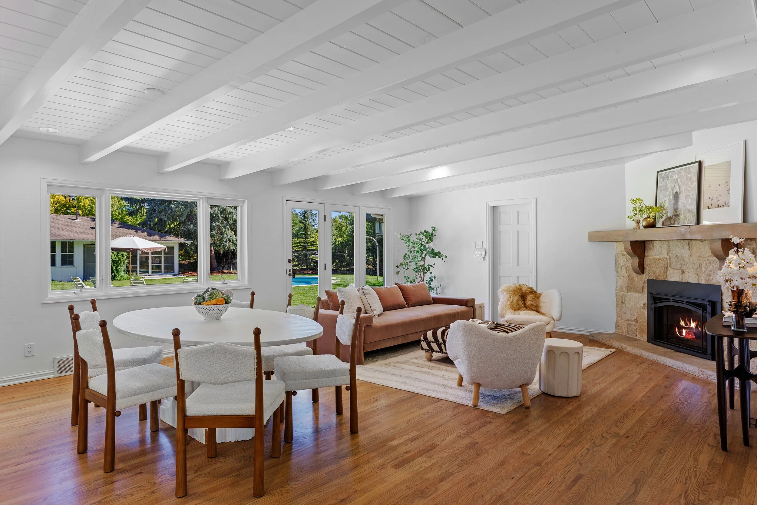 Bright living room with large windows, white walls, wood floors, a gas fireplace, and a mix of modern and vintage furniture.
