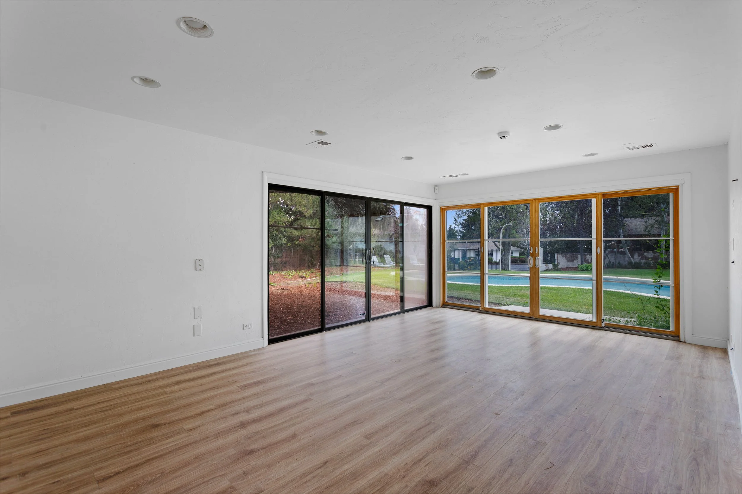 Empty living room with white walls, light wood flooring, and large sliding glass doors leading to a backyard with trees and a pool.
