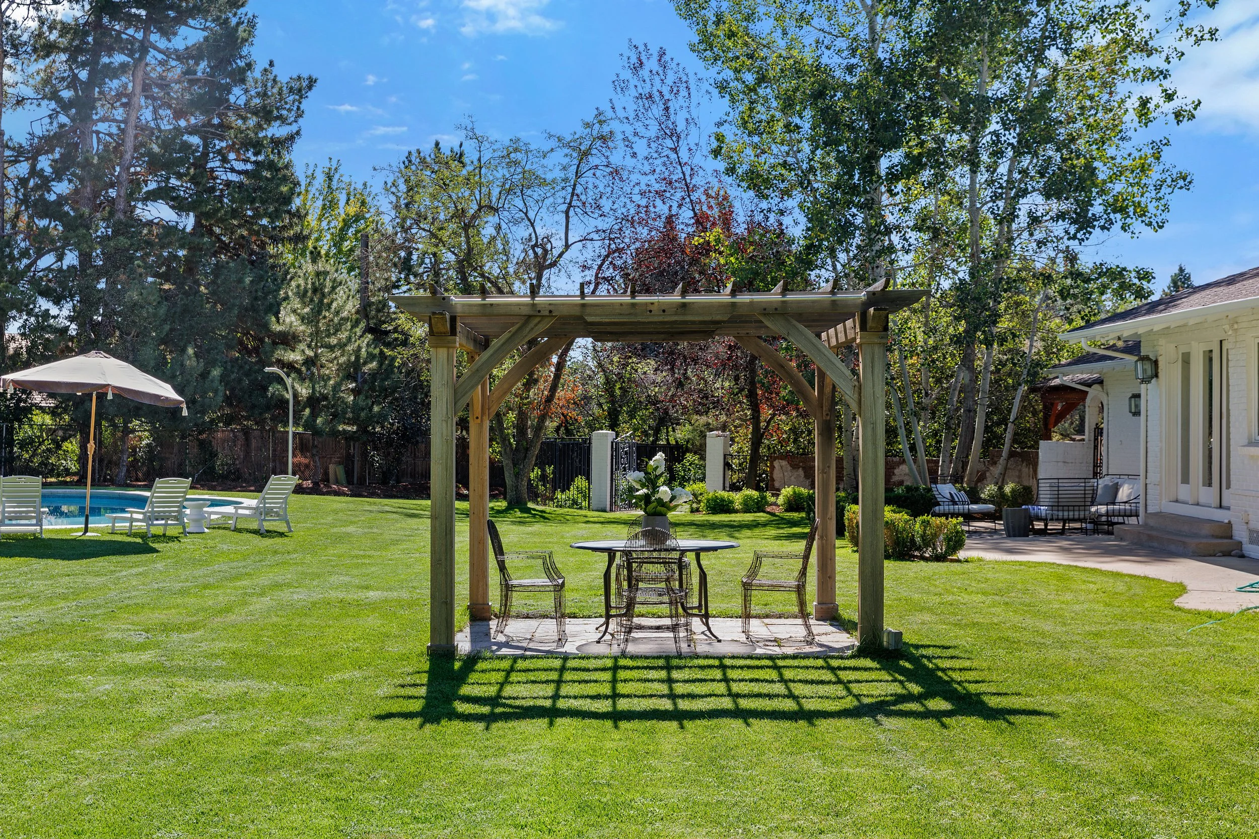 A backyard with a grassy lawn, a swimming pool with lounge chairs and an umbrella, a wooden pergola with a table and chairs, surrounded by trees and a house on the right.
