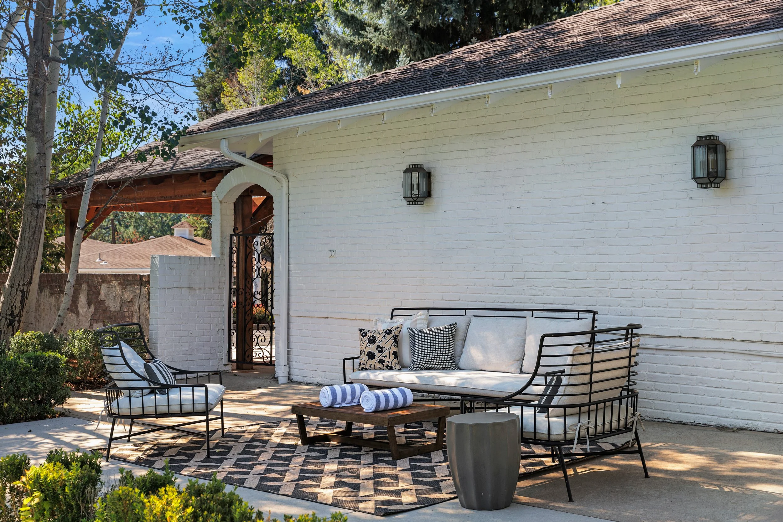 Outdoor patio area with black metal seating, white cushions, rolled towels, and a patterned rug, next to a white brick wall with black wall-mounted lanterns, greenery, and trees in the background.
