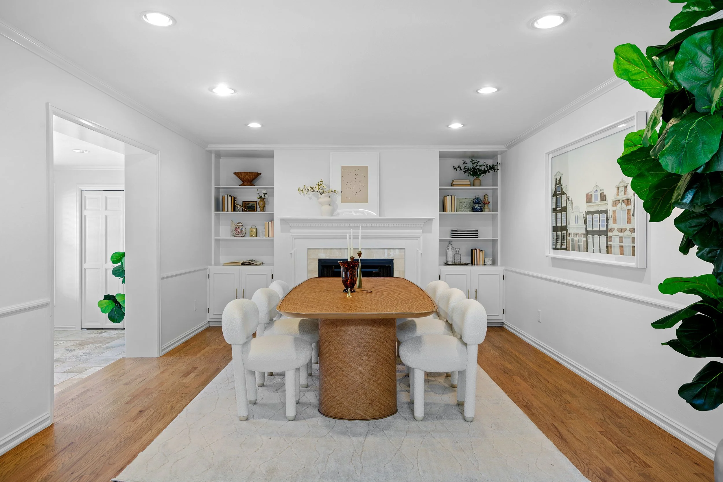 Bright dining room with a wooden table surrounded by eight white upholstered chairs, a beige side by side rug, white built-in shelves with decorative items, and a white fireplace, with large windows and green plants in the corners.
