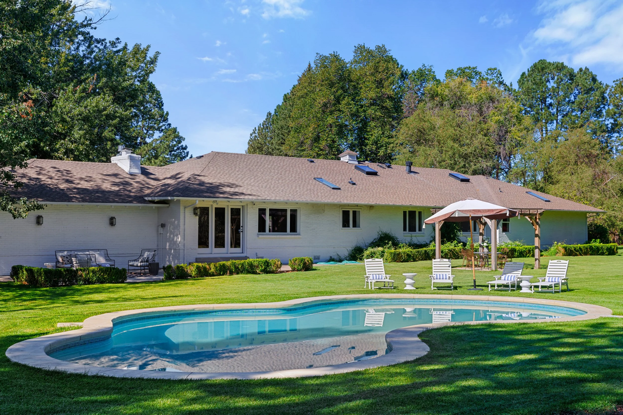 Backyard with a swimming pool, lawn chairs, patio table with an umbrella, and the back of a house with a patio. Trees and a blue sky in the background.