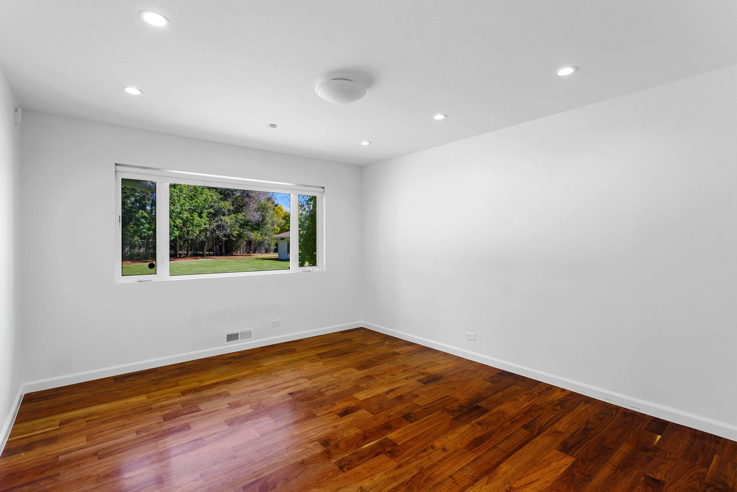Empty room with white walls, wood flooring, a large window showing green trees outside, and ceiling lights.