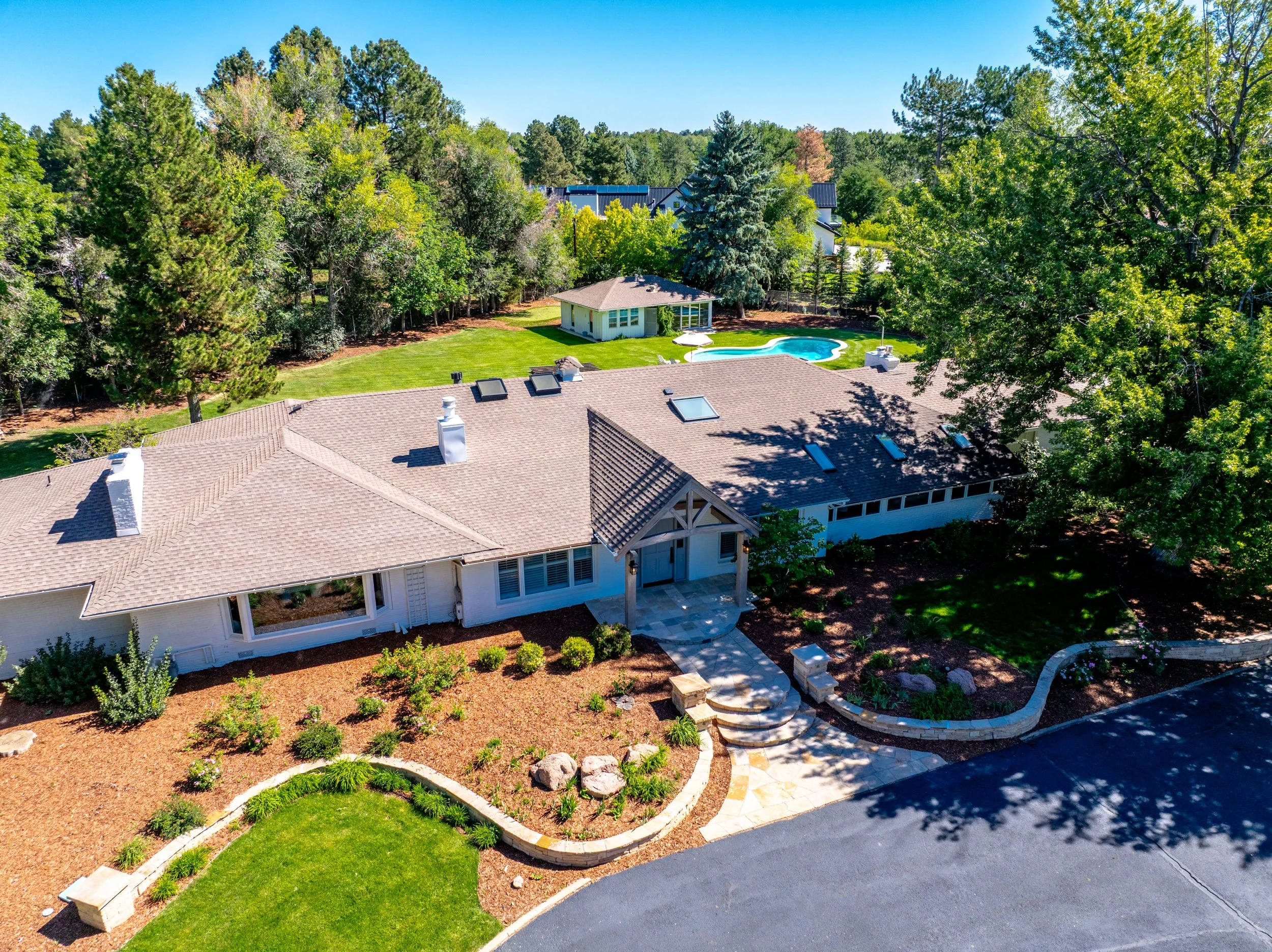 A suburban backyard featuring a swimming pool, a small pool house, a landscaped garden with mulch and plants, a paved pathway with steps, and a neighboring house surrounded by trees under a clear blue sky.