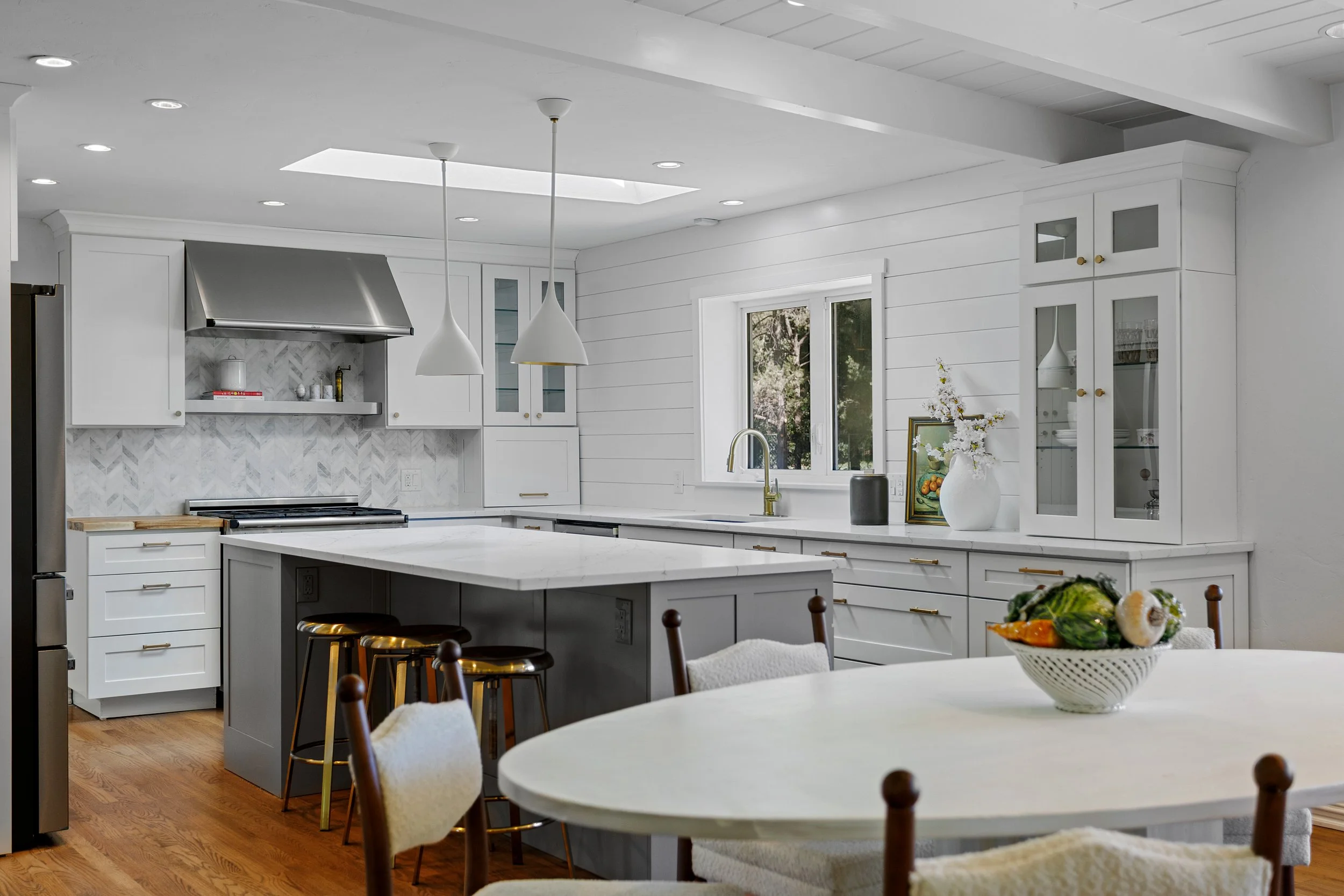 A bright, modern white kitchen with a large kitchen island, pendant lights, and white cabinetry. There is a window above the sink with a view of trees outside, and a fruit bowl on a round dining table in the foreground.