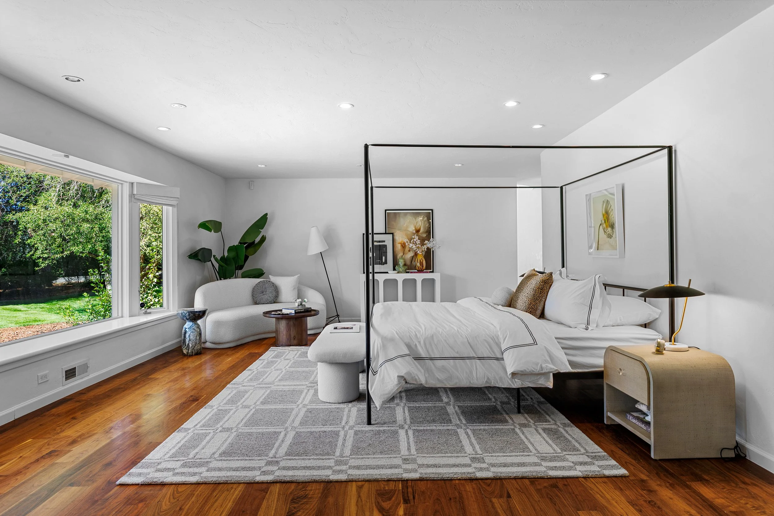 A bright, modern bedroom with a large window, white walls, and hardwood floors. The room features a white bed with black trim, a side table, a white lounge chair with pillows, and framed artwork on the wall.