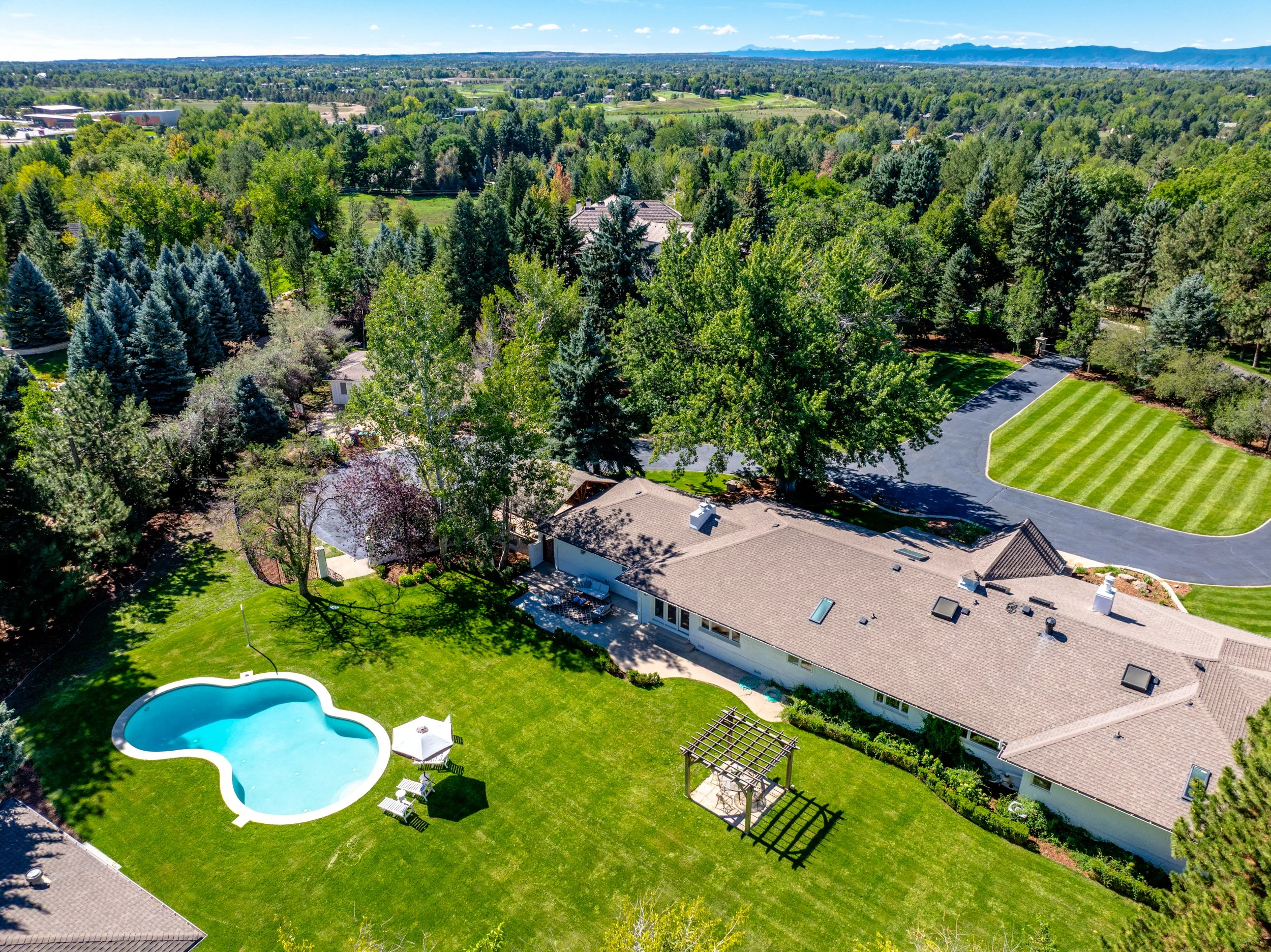 Aerial view of a backyard with a kidney-shaped swimming pool, a grassy lawn, a patio with lounge chairs and a small gazebo. Surrounded by trees and neighboring houses, with distant mountains in the background.