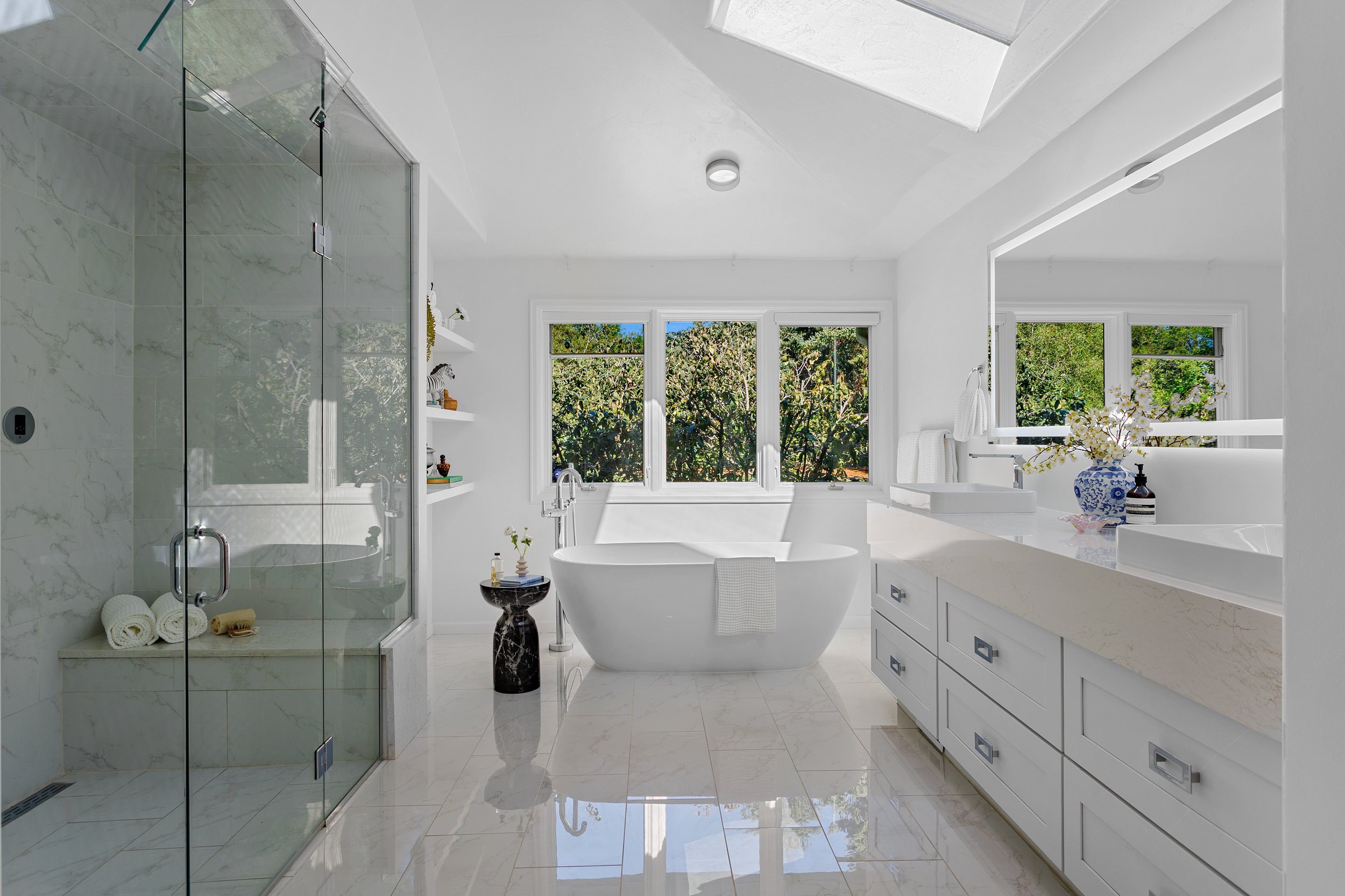 Modern white bathroom with a glass-enclosed shower, a freestanding bathtub, and a vanity with dual sinks, large windows with greenery outside, and decorative items on the counter.