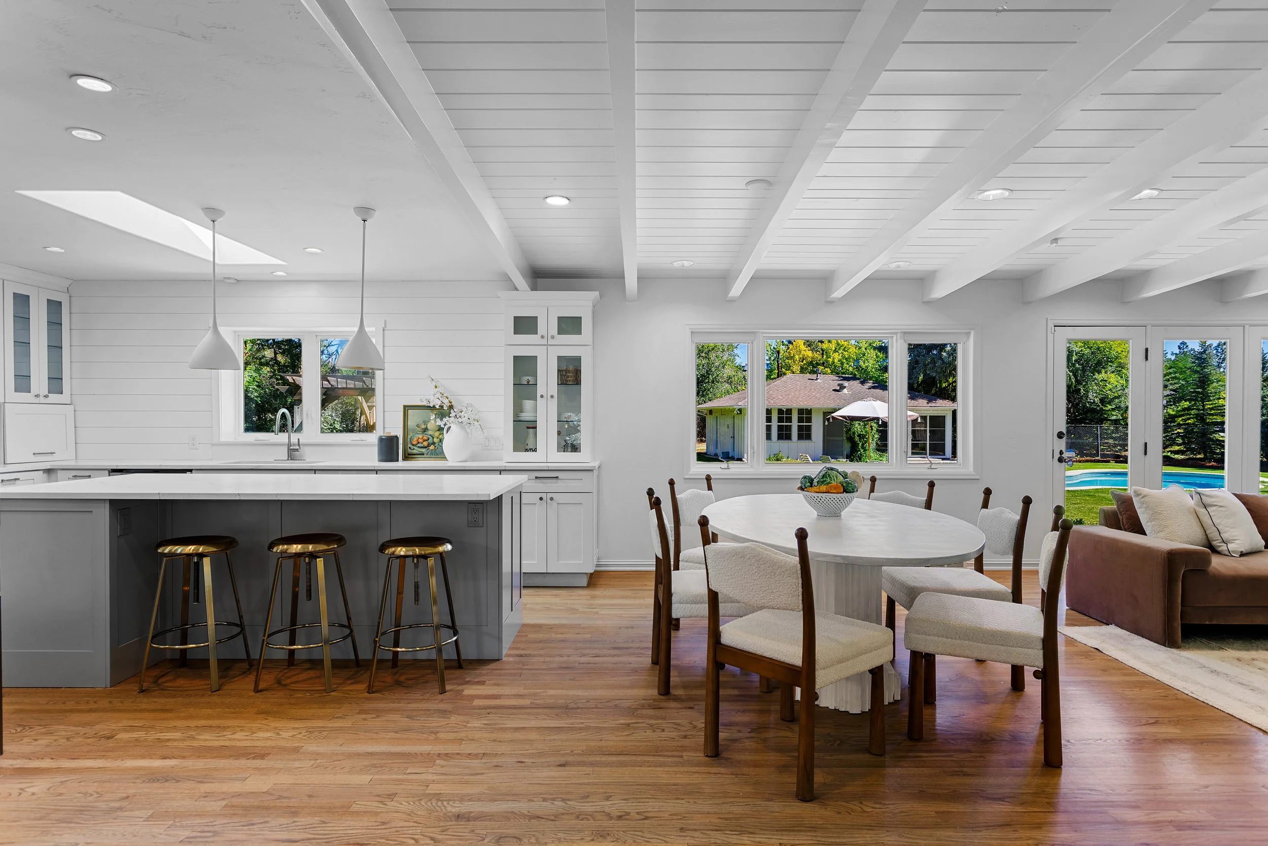 Bright open plan kitchen and dining area with white cabinetry, a large island with barstools, and a round dining table with chairs. Large windows and a glass door reveal a backyard with trees, a house, and a swimming pool. Ceiling with exposed beams 