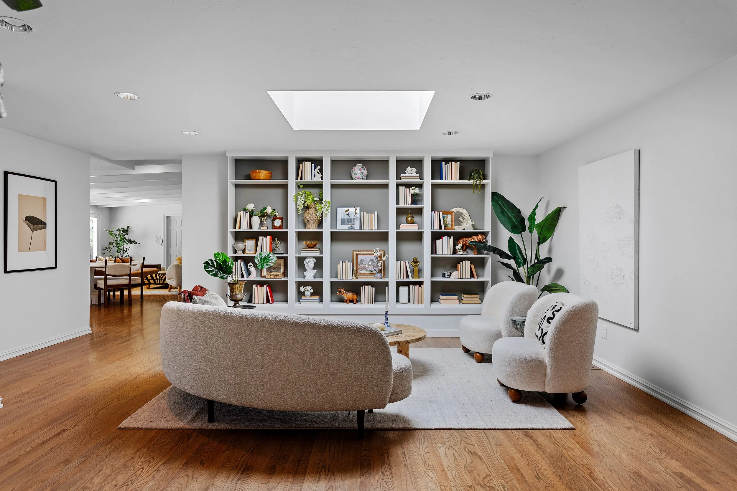 A living room with white walls, hardwood floors, white upholstered furniture, and a large built-in bookshelf filled with various decorative items and books. There is a doorway leading to a dining area with a wooden table and chairs. Potted plants are