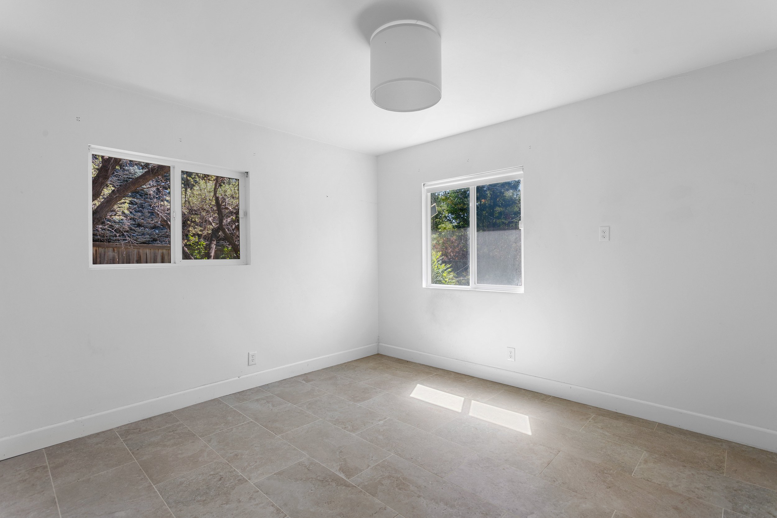 Empty room with white walls, two windows showing trees outside, tiled floor, and ceiling light fixture.