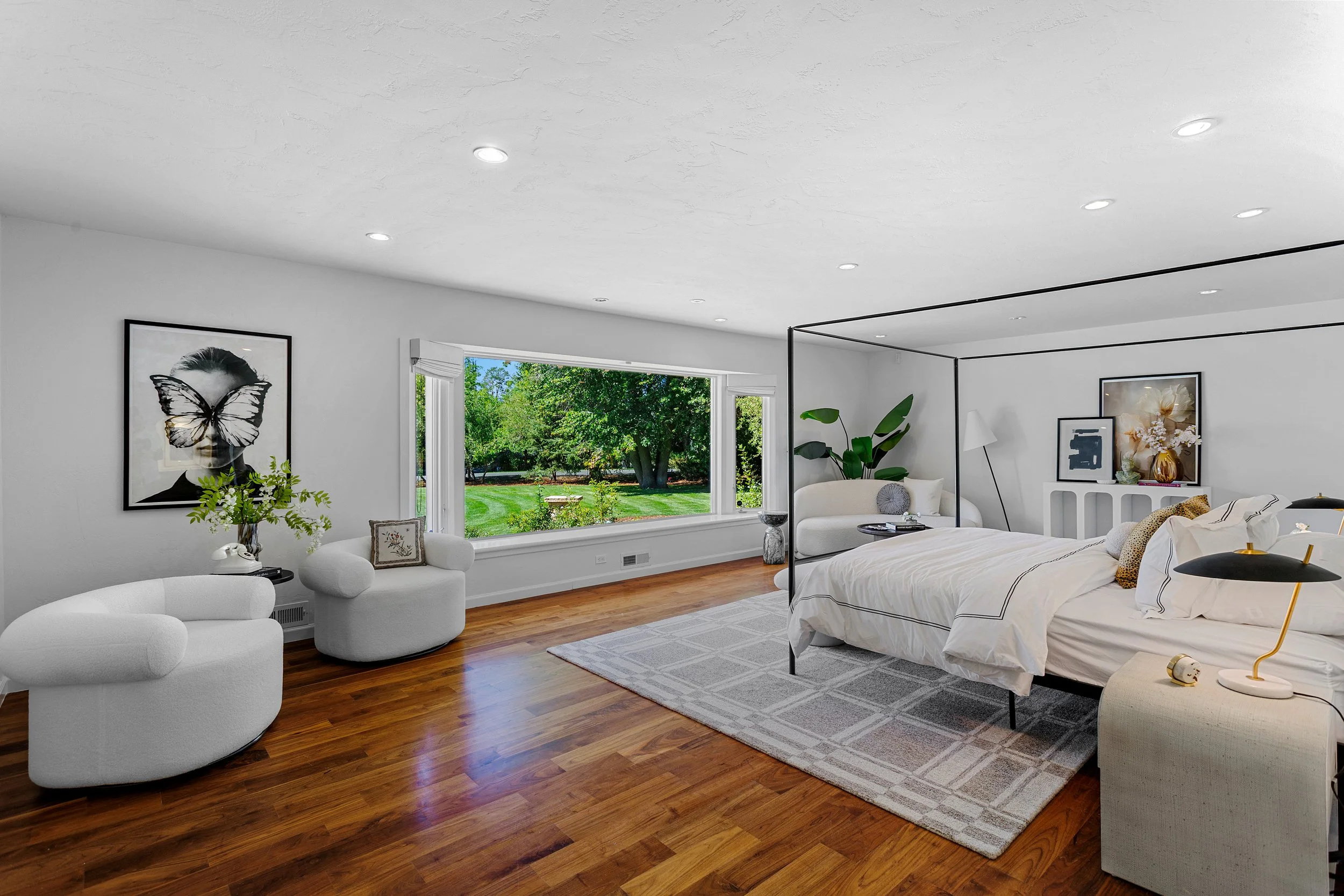 A bright bedroom with white walls, wooden floor, and large window showing a green outdoor area. The room has a white bed with black piping, a white armchair, framed artwork including a portrait with a butterfly mask, a white side table, and decorativ