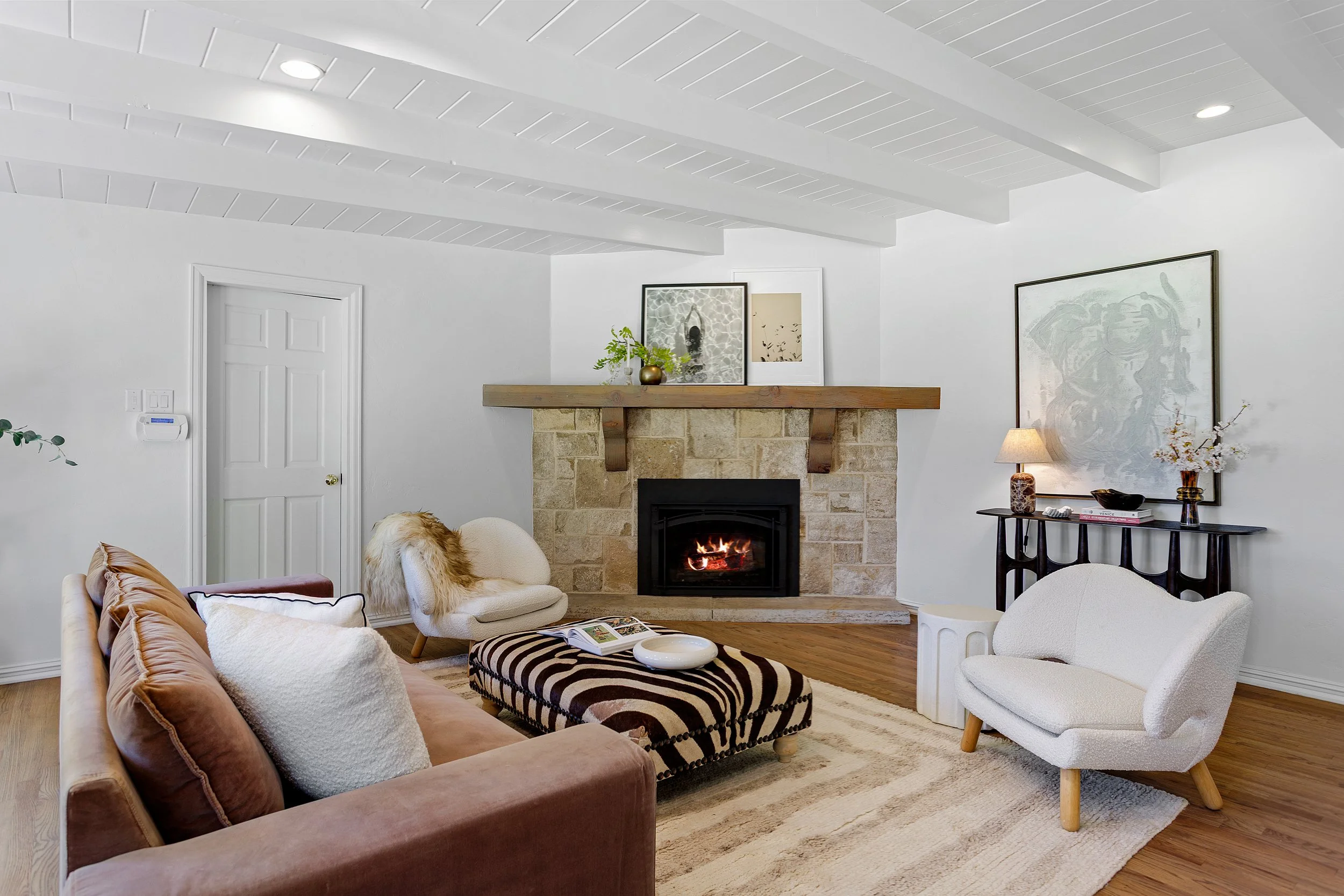 A living room with a beige couch and a white armchair arranged around a zebra-print ottoman in front of a fireplace with stone surround and a wooden mantel. The walls are white with framed artwork, and a small table with a lamp and decorative items i