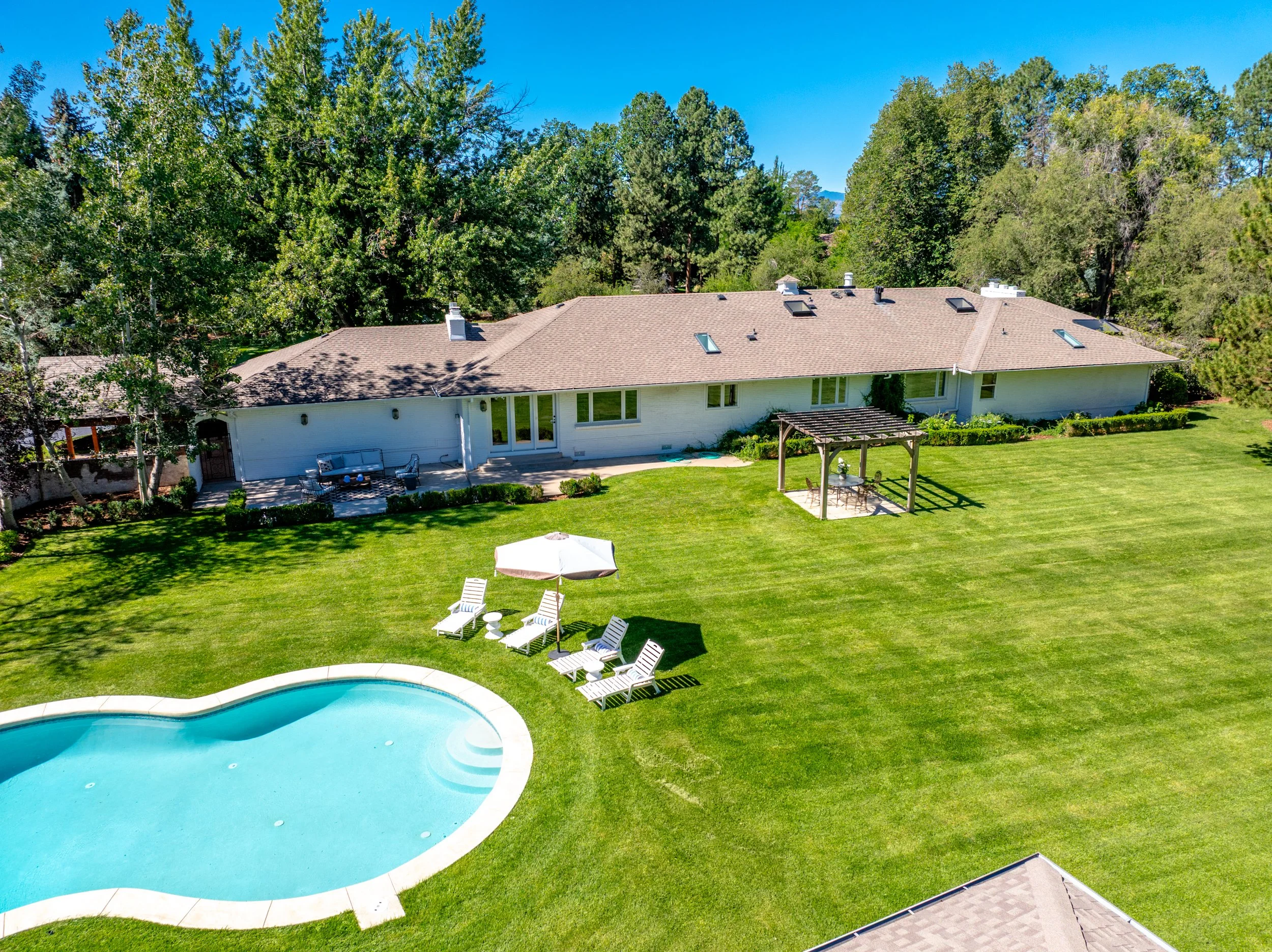 A backyard with a swimming pool, white lounge chairs, a patio table with chairs and umbrella, surrounded by a green lawn and trees, with a house in the background and a bright blue sky.