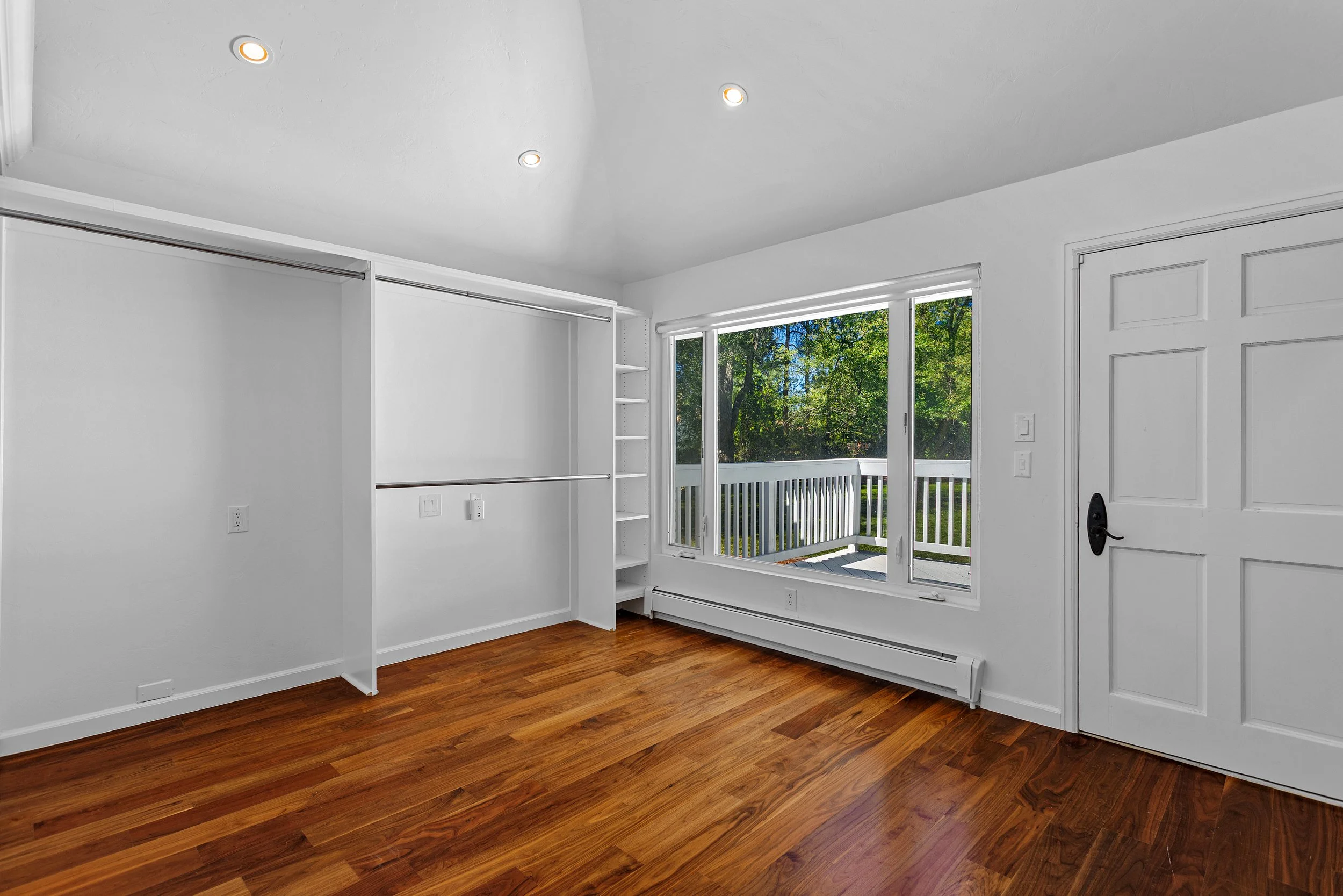 Empty room with hardwood floors, white walls, large window showing trees outside, and other built-in features.
