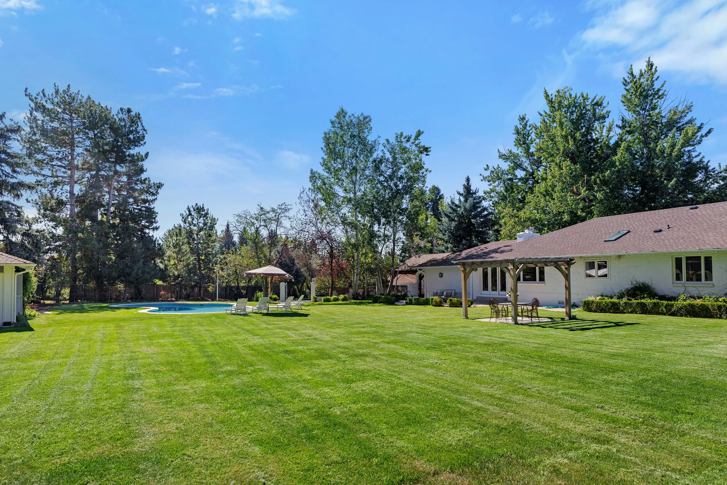 Backyard with well-manicured green lawn, outdoor swimming pool surrounded by lounge chairs, umbrellas, a white house with a brown roof, patio furniture, and tall trees under a bright blue sky with some clouds.