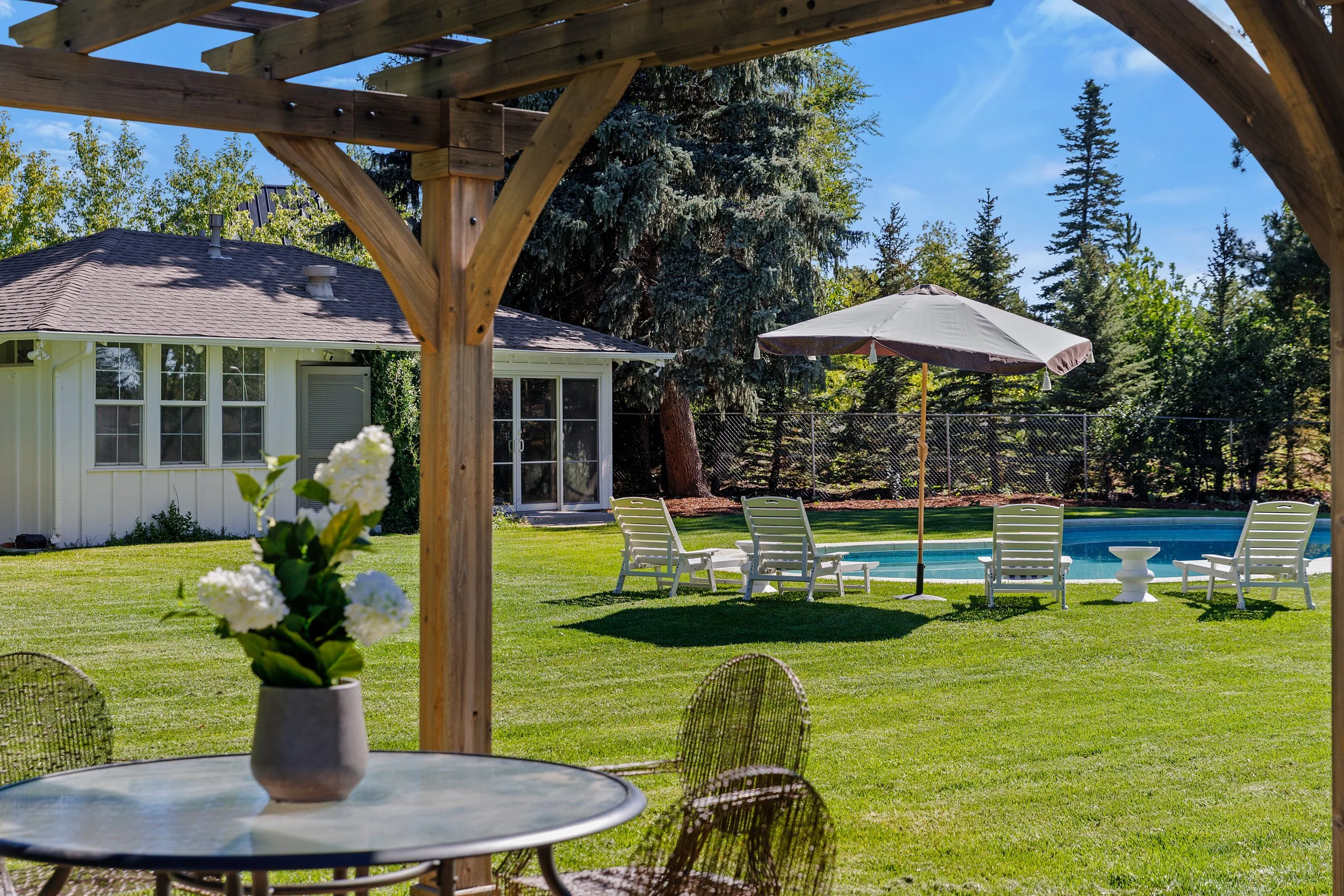 A backyard with a swimming pool, outdoor chairs, a large umbrella, a house with a sliding glass door, tall pine trees, and a pergola structure in the foreground with a flower vase on a table.