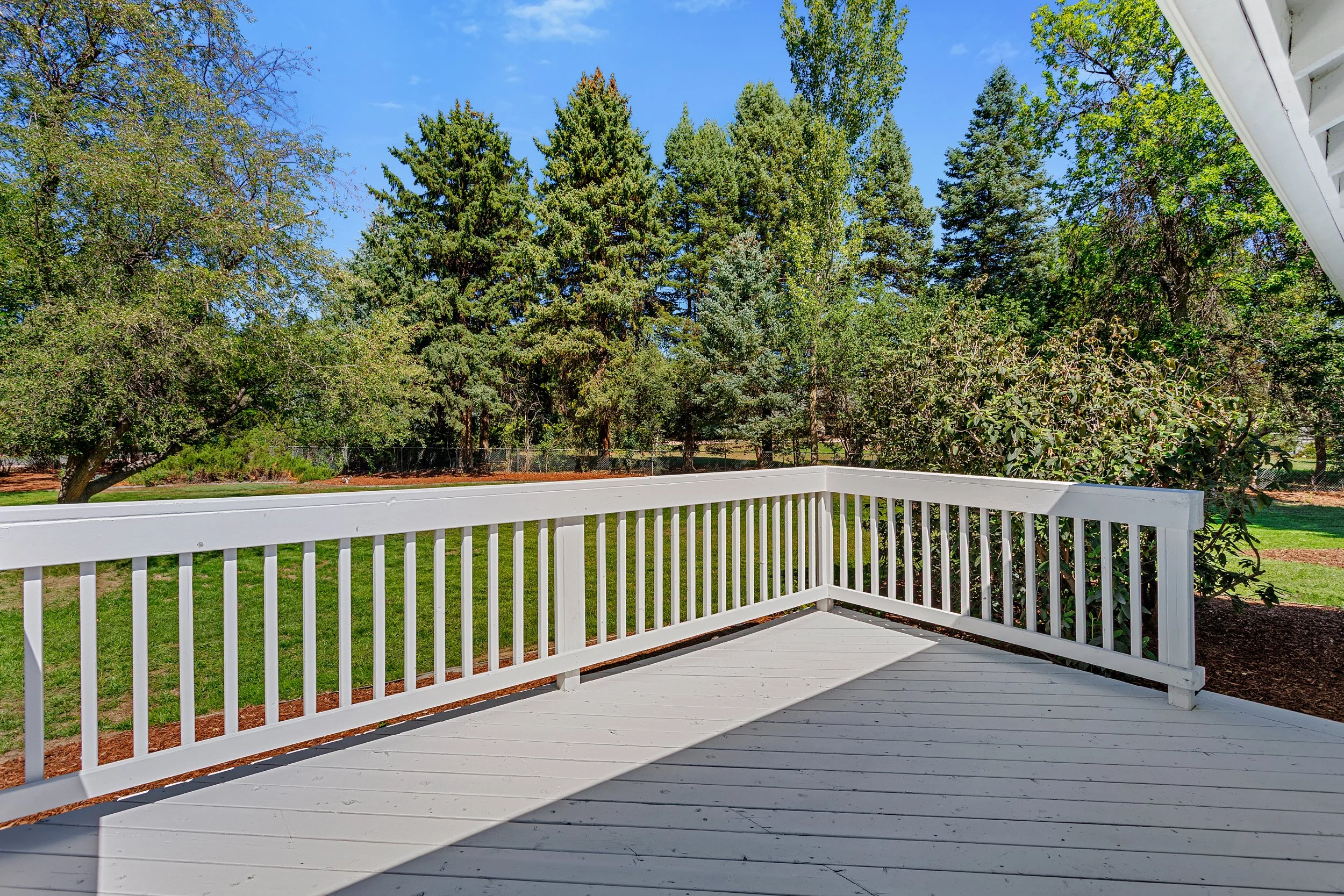 View from a white wooden deck with a white railing overlooking a grassy yard and a wooded area with green trees under a clear blue sky.