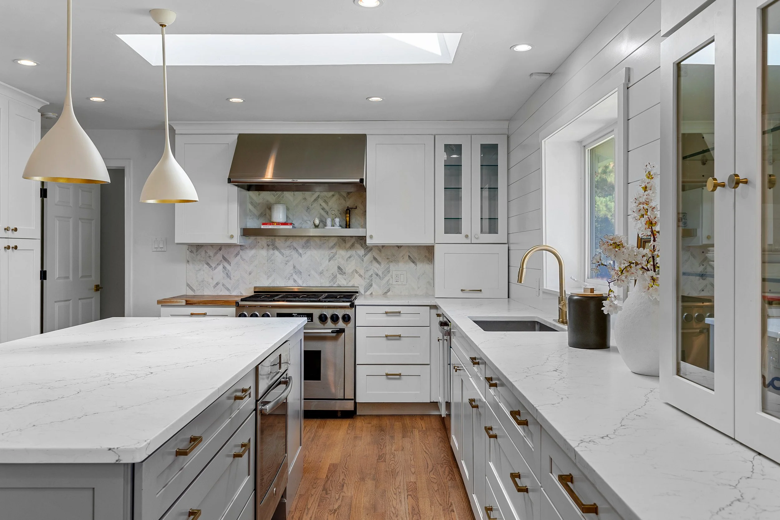 Modern white kitchen with marble countertops, wooden flooring, stainless steel stove, glass cabinets, and a large skylight.