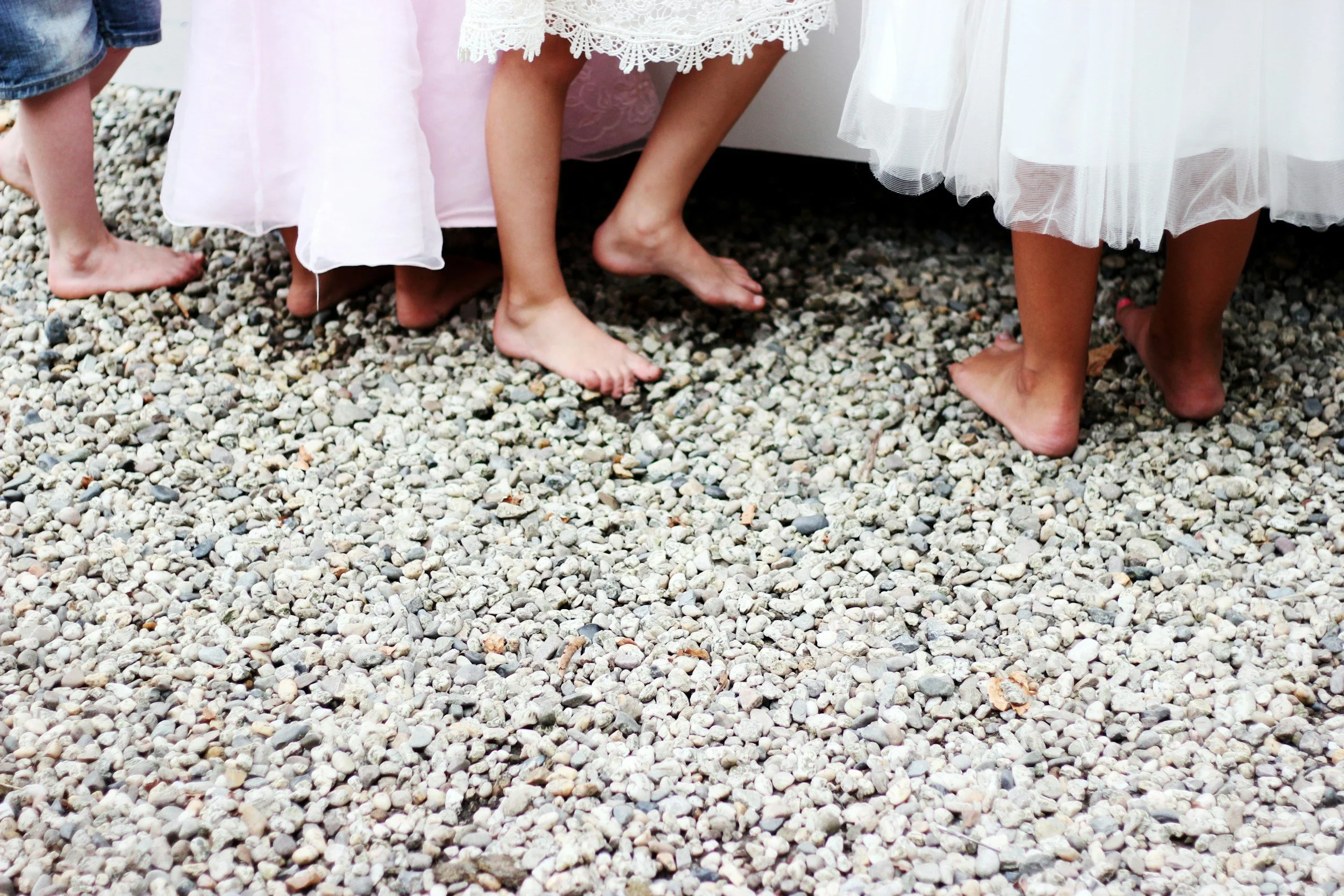A group of children dressed in fancy dresses and shorts, barefoot on a gravel surface, with some children partially visible.