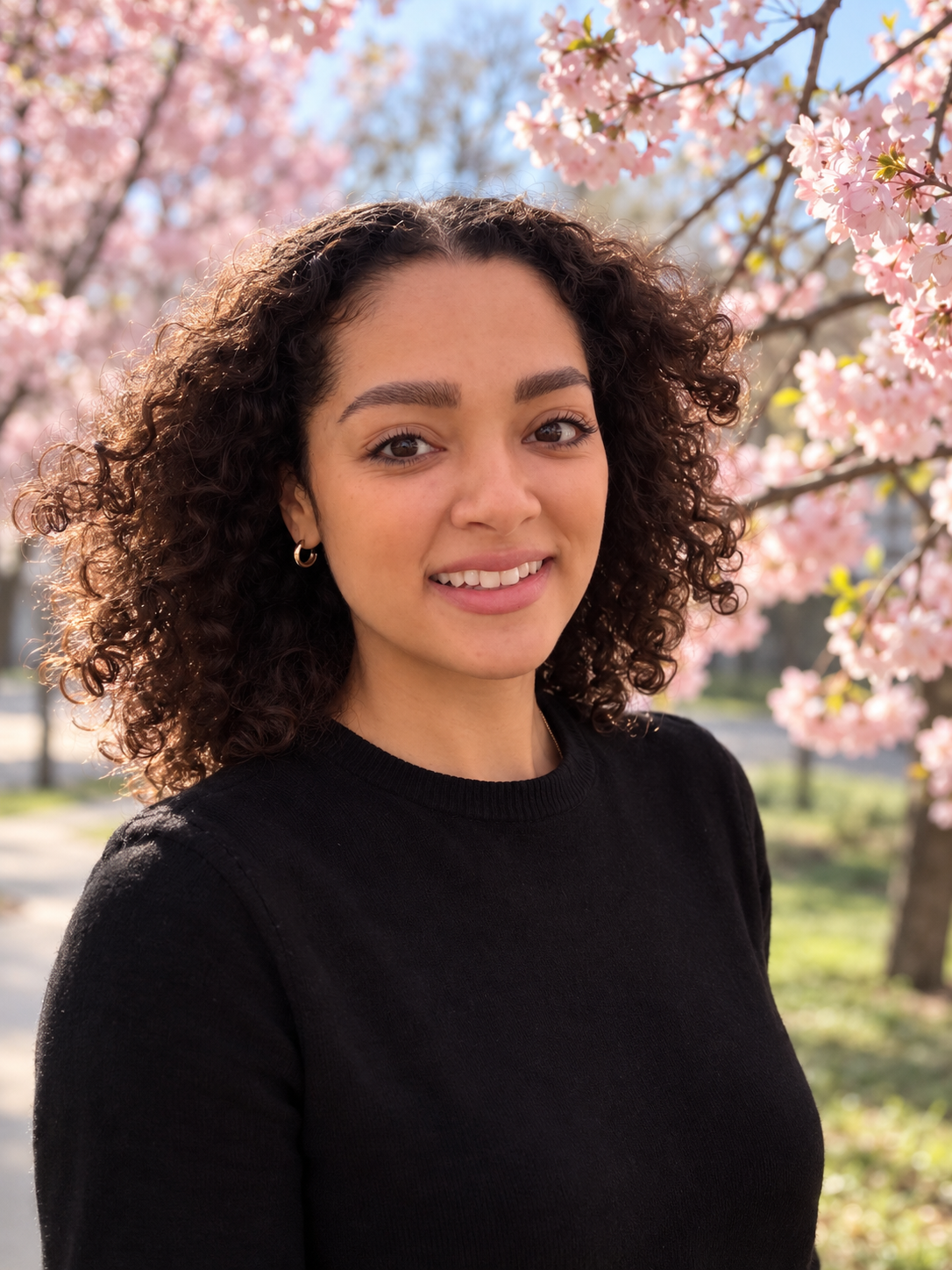 A woman with curly brown hair, wearing a black top, standing outdoors in front of pink cherry blossom trees during spring.