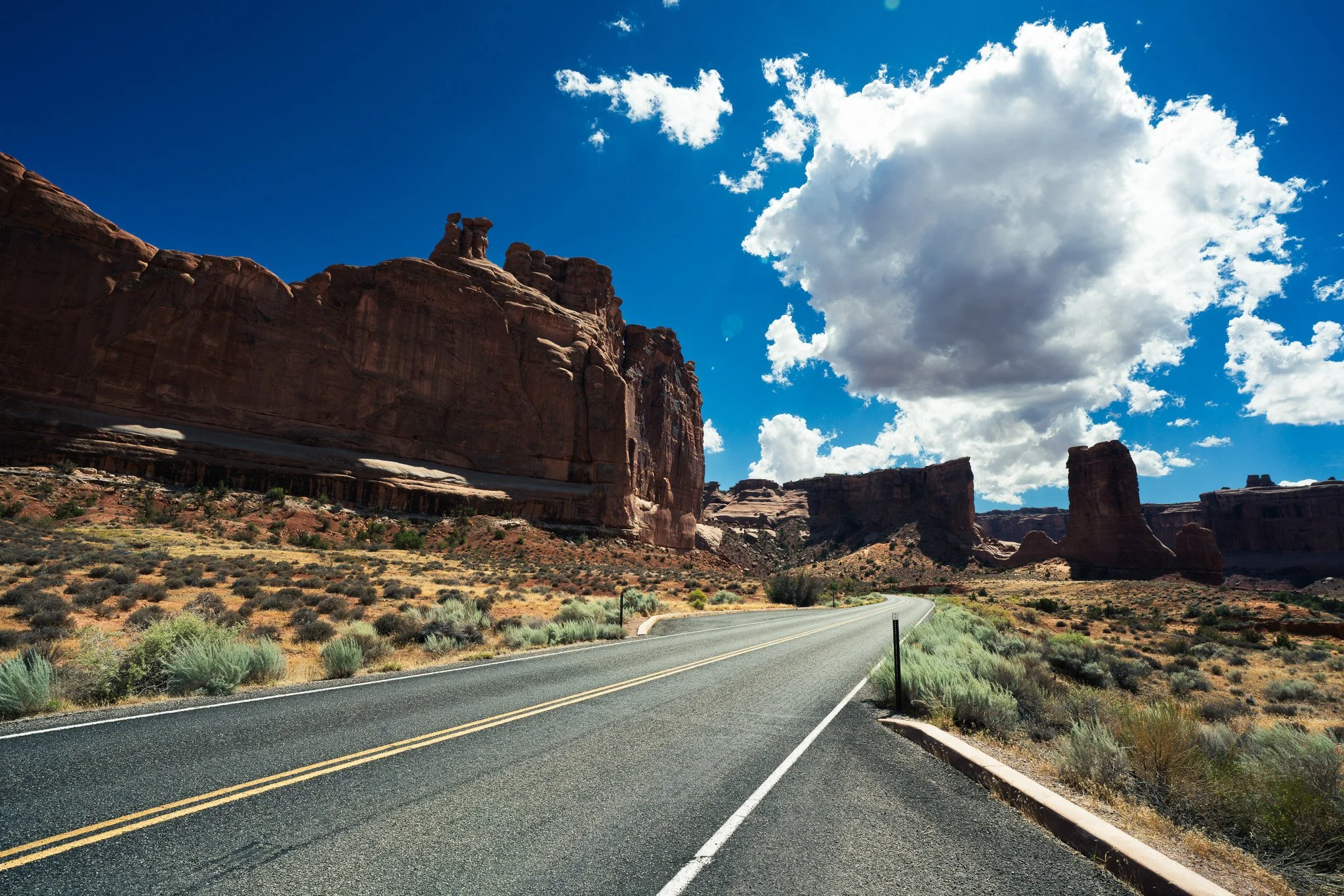 A winding two-lane road runs through a desert landscape with large red rock formations on the left and sparse vegetation on the right. The sky above is bright blue with scattered white clouds.