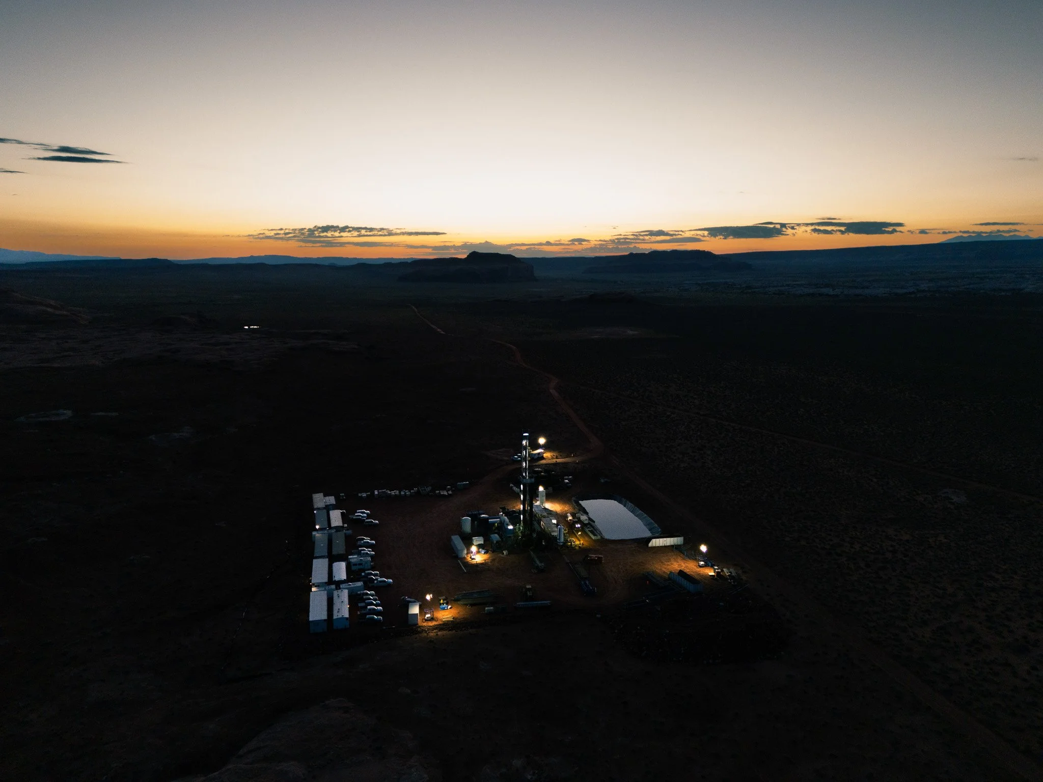 A landscape at dusk or dawn with a flat terrain, distant mountains, and a sky with a gradient from orange to pale blue. There are several buildings and parked vehicles illuminated in the foreground, indicating a settlement or facility in a remote are