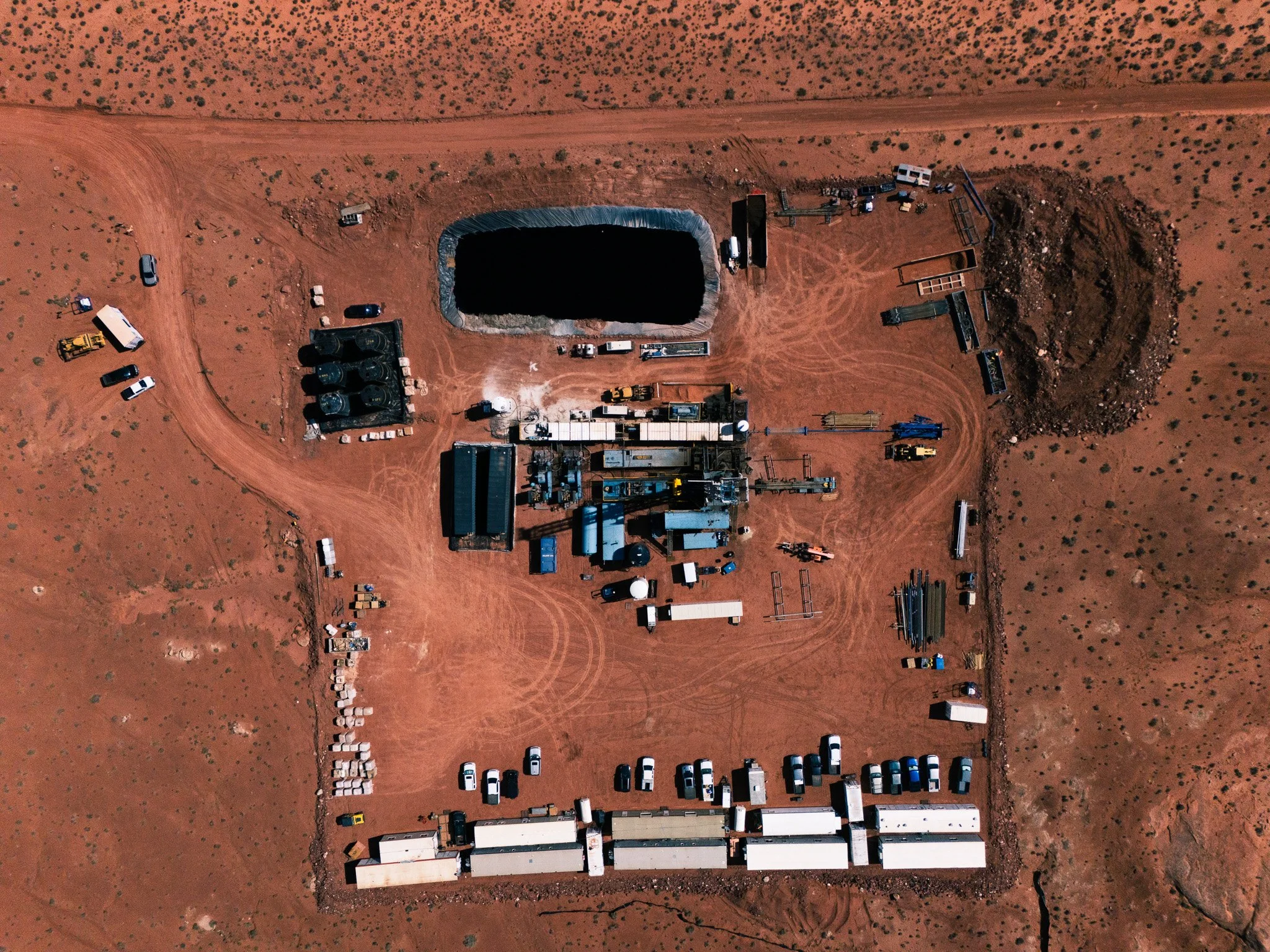 An aerial view of a construction site with a large rectangular pond surrounded by various construction equipment and vehicles on red dirt terrain.