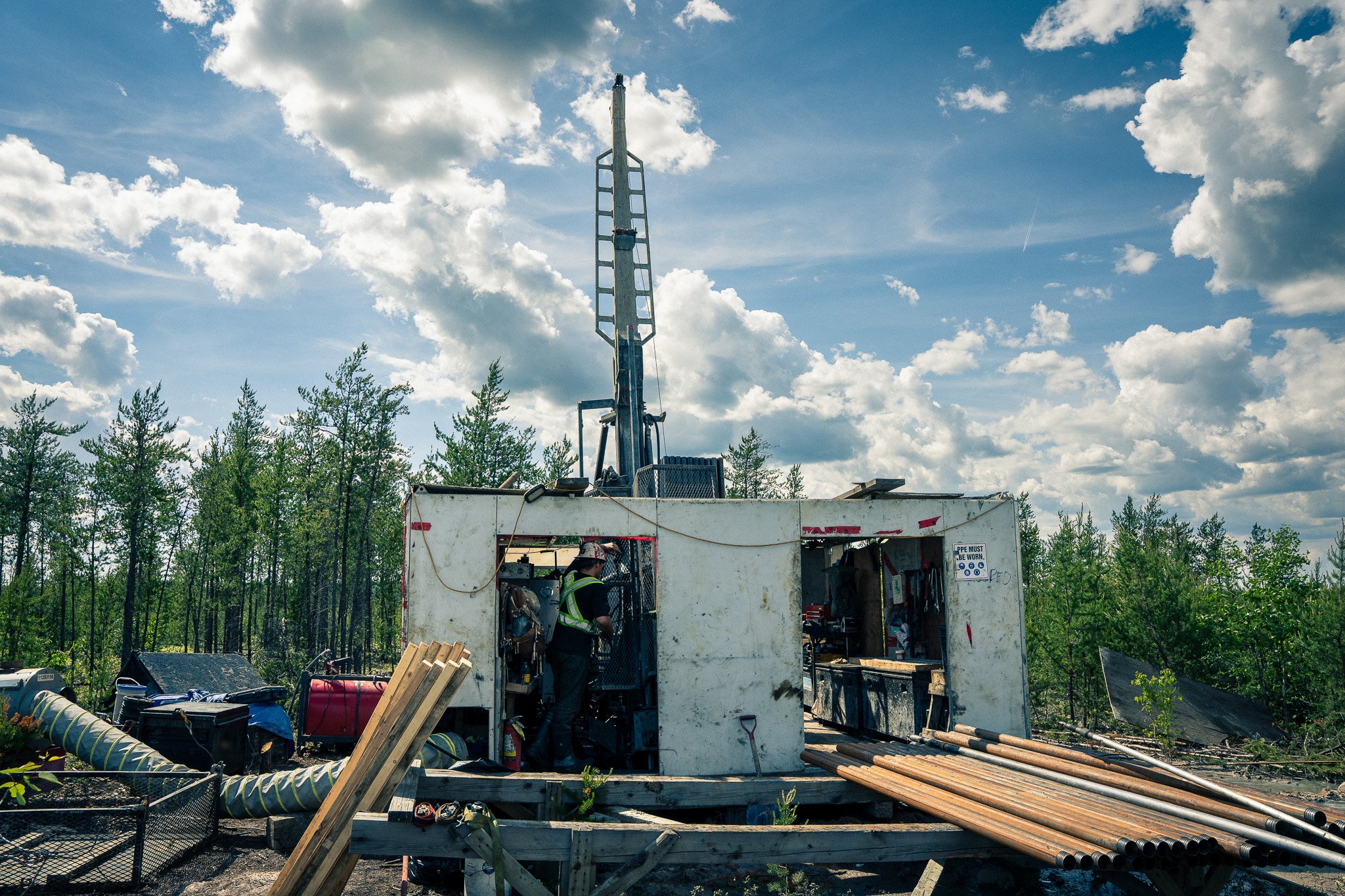 A worker preparing equipment inside a service truck with construction pipes and tools on the ground, trees and cloudy sky in the background.