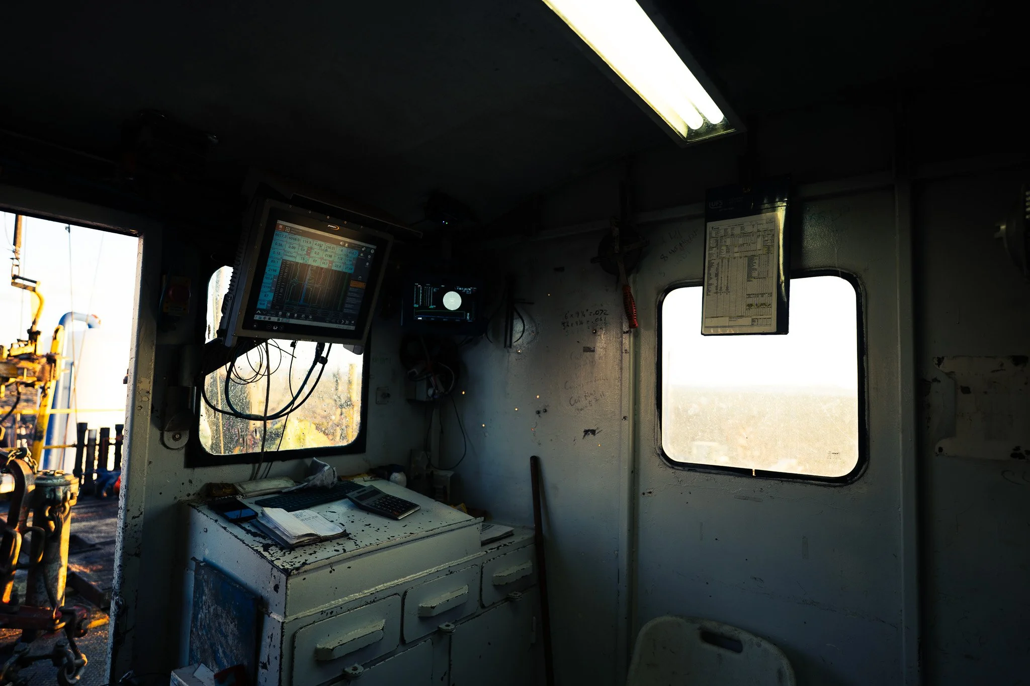 The interior of a control room with two monitors displaying various data, a white desk with papers and a calculator, and windows showing an outdoor industrial area with pipes and machinery.