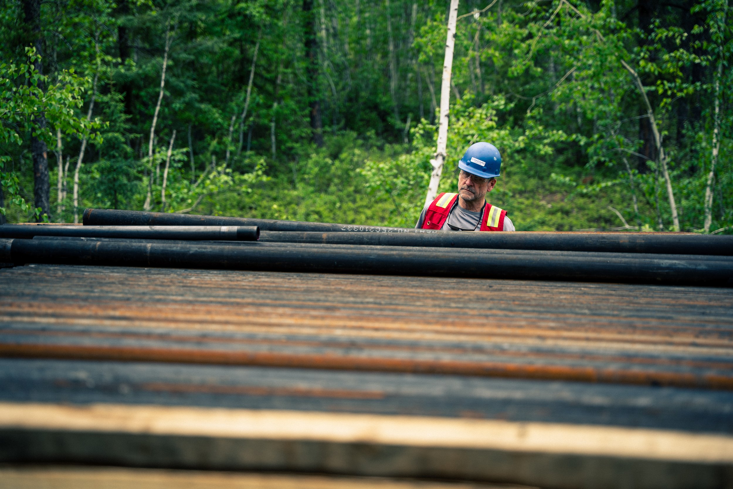 A man wearing a blue hard hat, safety glasses, a red safety vest, and gray shirt standing outdoors in a lush green forest surrounded by trees, behind a stack of black pipes.