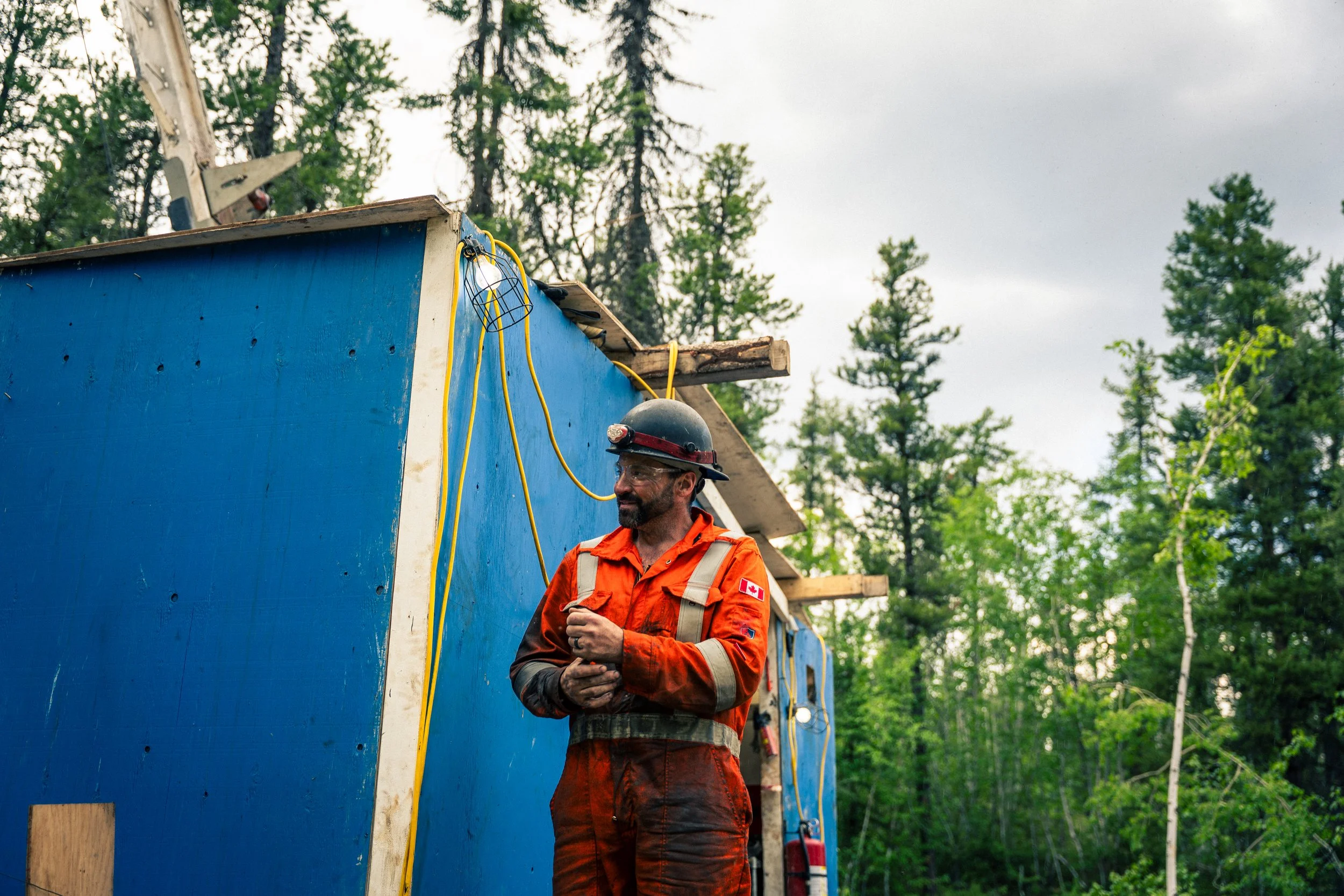 A construction worker in an orange safety jacket and a helmet stands next to a blue wall under construction outdoors, with trees in the background.