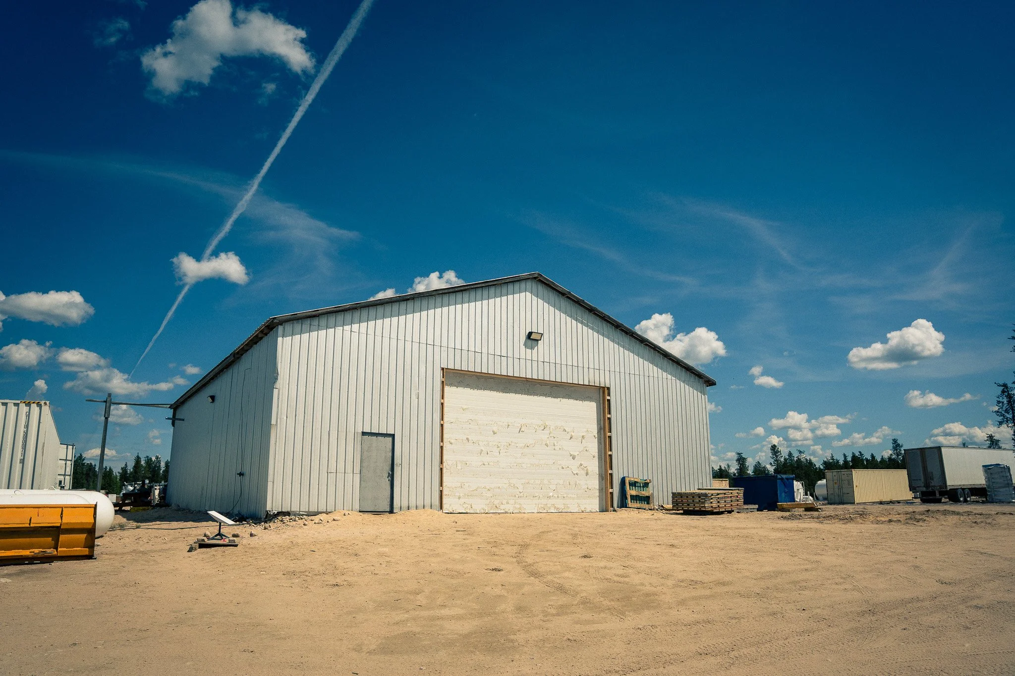 An industrial warehouse building with a metal exterior, set against a bright blue sky with scattered clouds and contrails. The area around the building is a dirt lot with various equipment and materials.