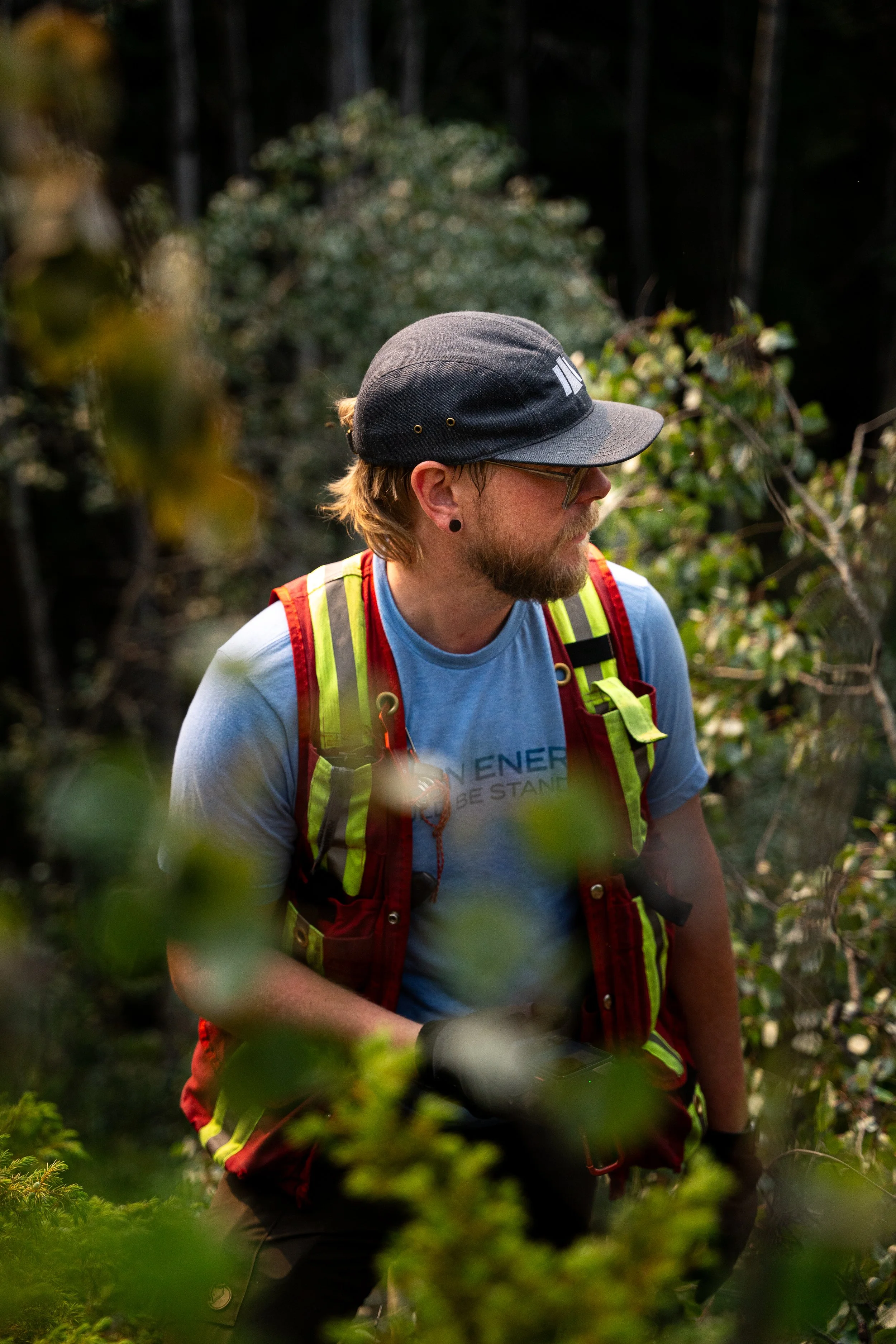 A young man with long hair, a beard, glasses, and a black cap, dressed in a blue t-shirt and a red backpack, exploring a forest trail during dusk with dense trees and foliage around him.