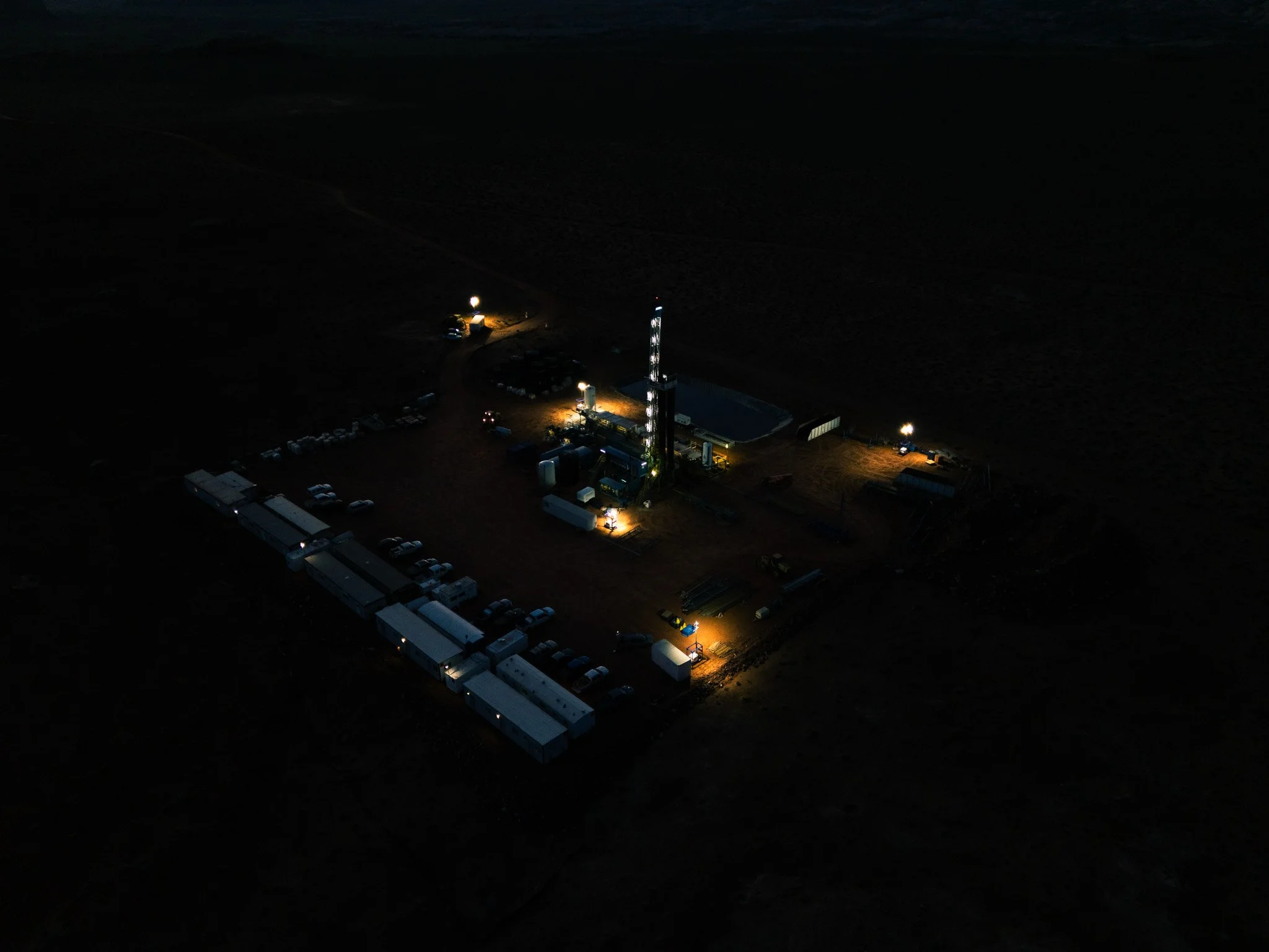 Aerial nighttime view of an industrial site with illuminated structures and parked vehicles, surrounded by darkness.