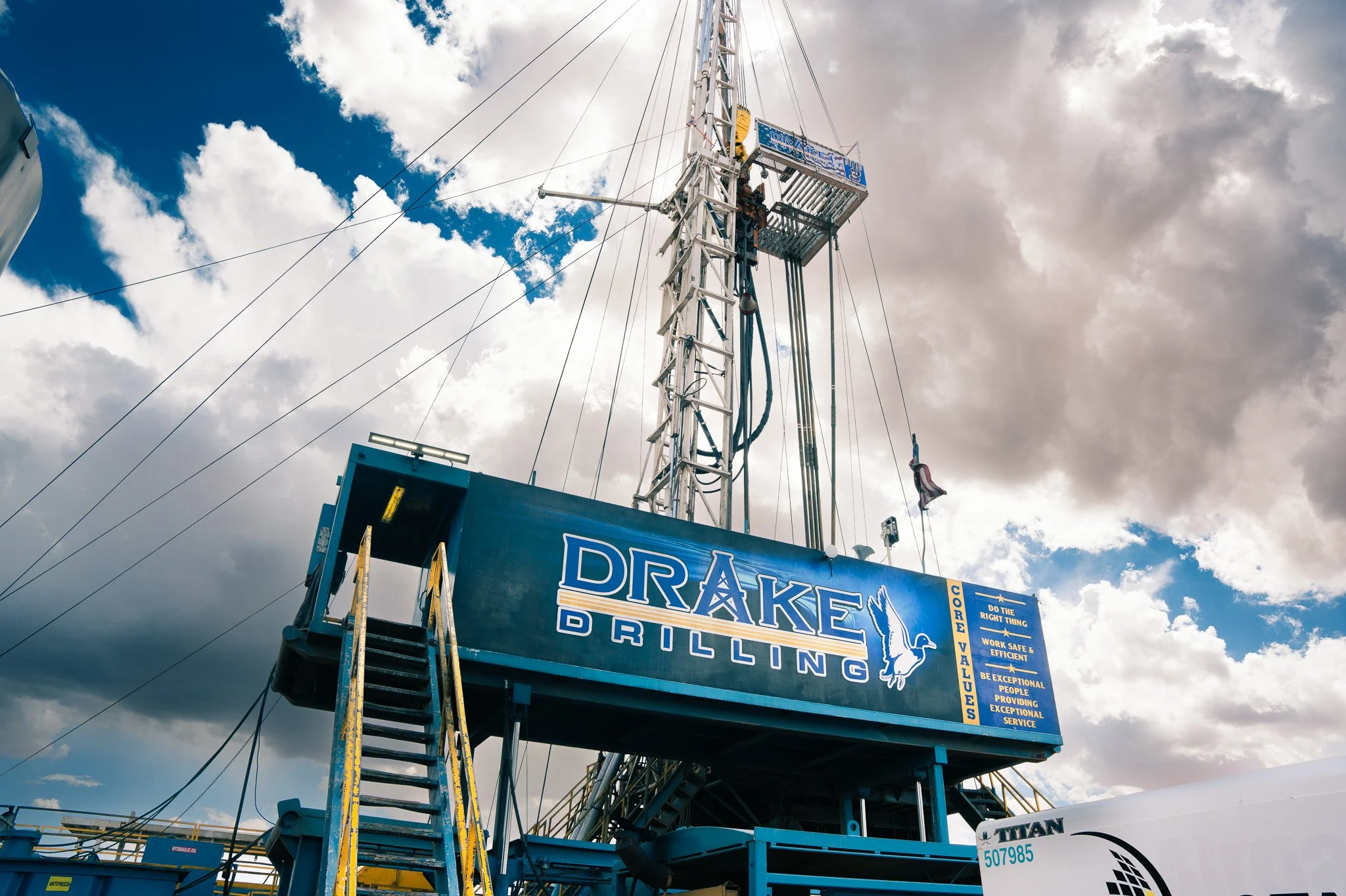 A drilling rig structure with a ladder, a worker working on the platform, and a sign that reads 'Drake Drilling' against a cloudy sky.