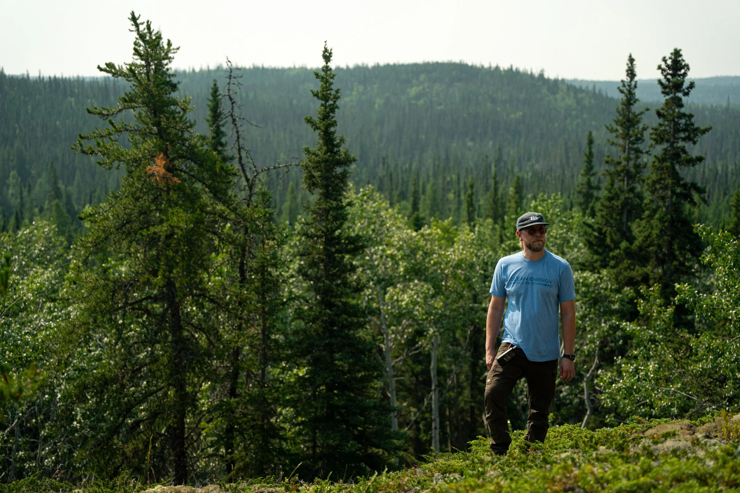 A man with a beard wearing sunglasses, a light blue t-shirt, and khaki pants, walking through a lush green forest with tall pine trees and mountains in the background.
