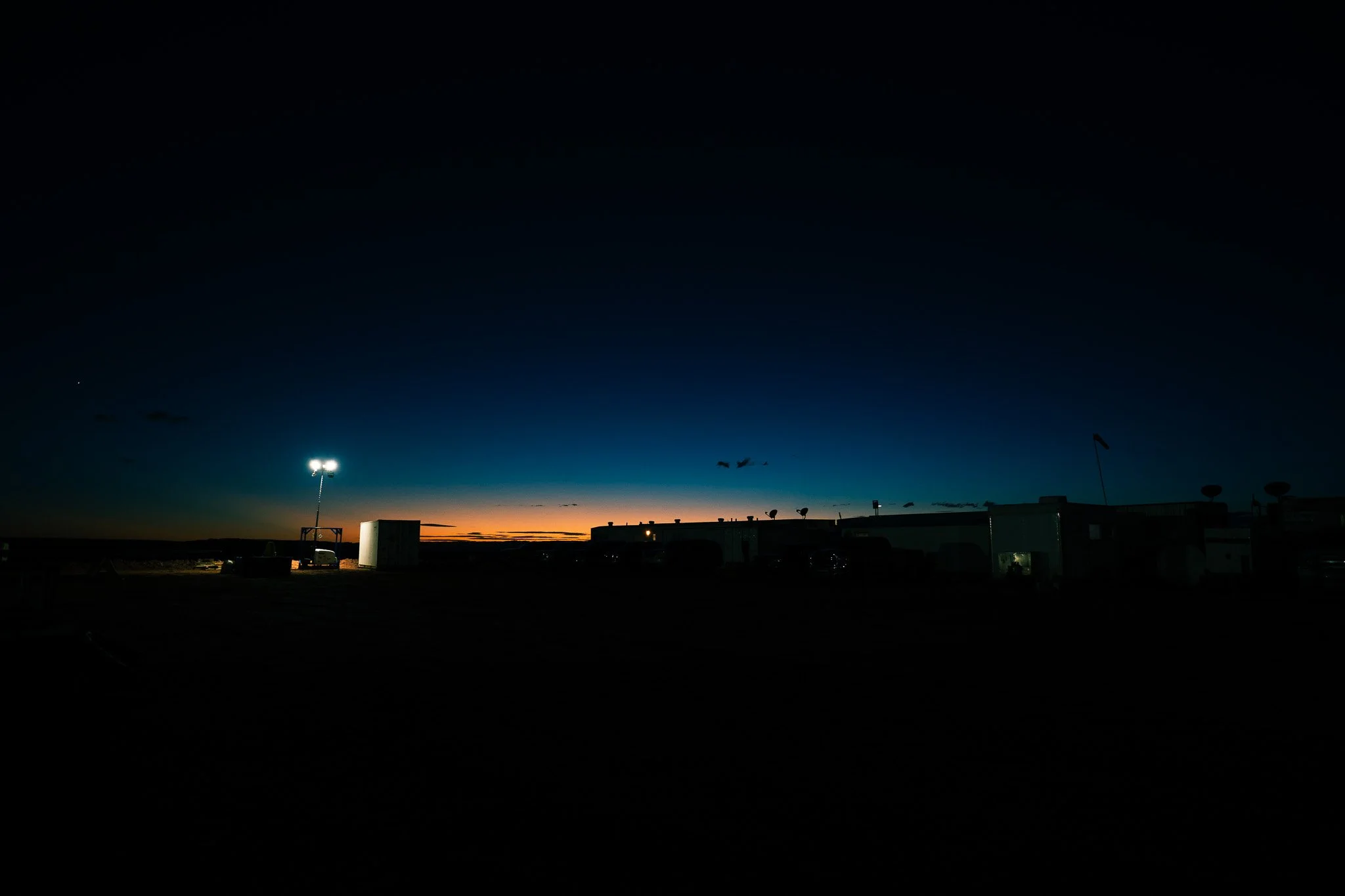 Nighttime view of a dark sky with a faint sunset on the horizon, silhouetted buildings, and bright stadium lights illuminating the area.