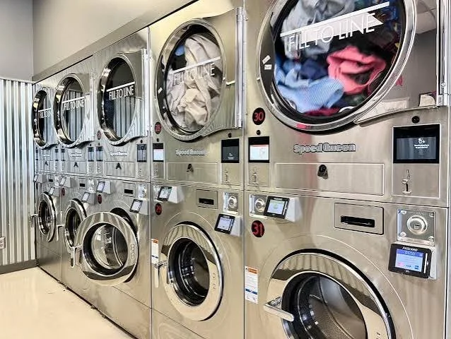 Multiple industrial stainless steel washing machines in a laundromat, filled with clothes.