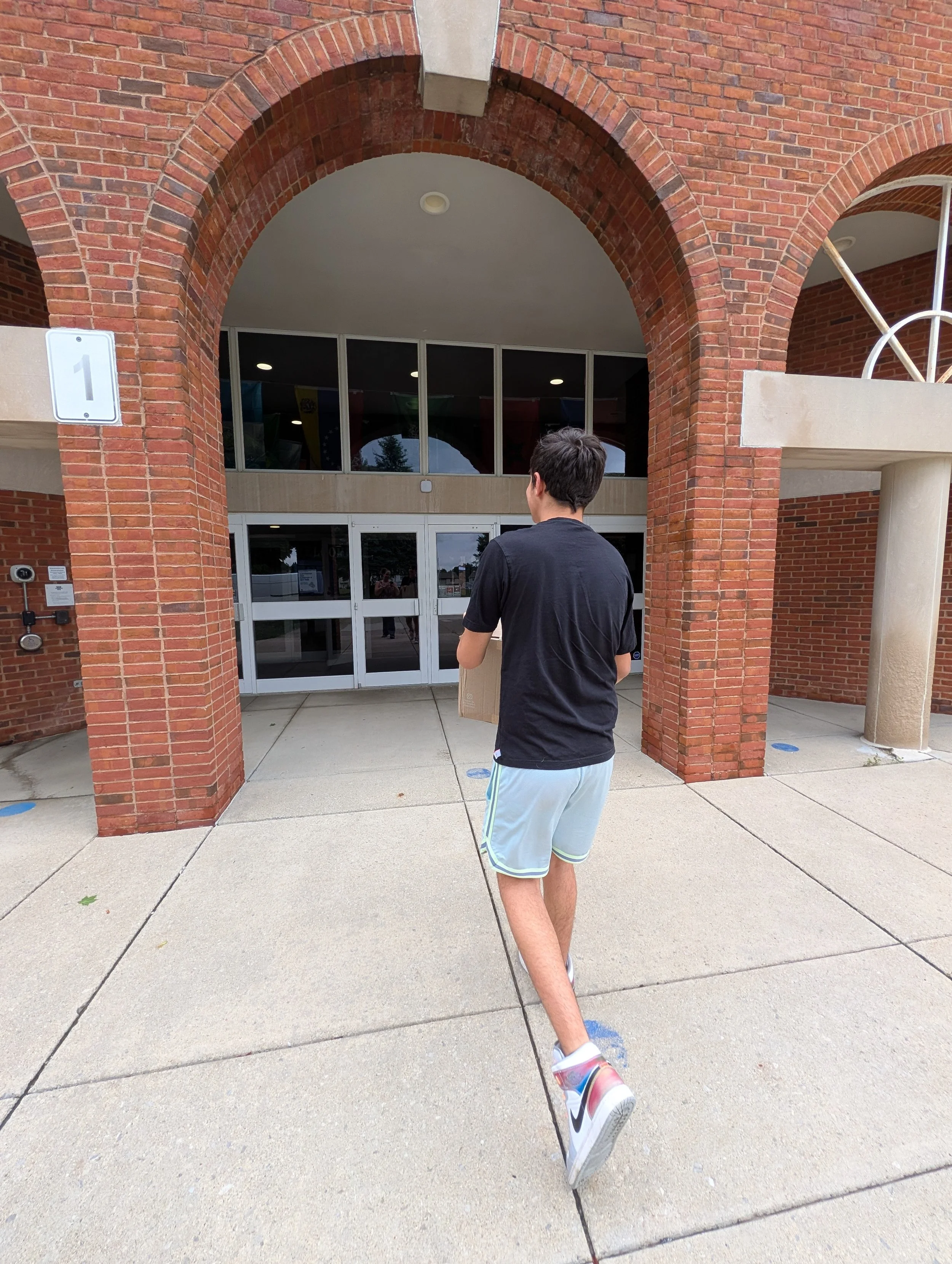A young man wearing a black t-shirt and light blue shorts walking away from the camera on a concrete sidewalk outside a brick building with large glass doors and windows. He is holding a cardboard box.