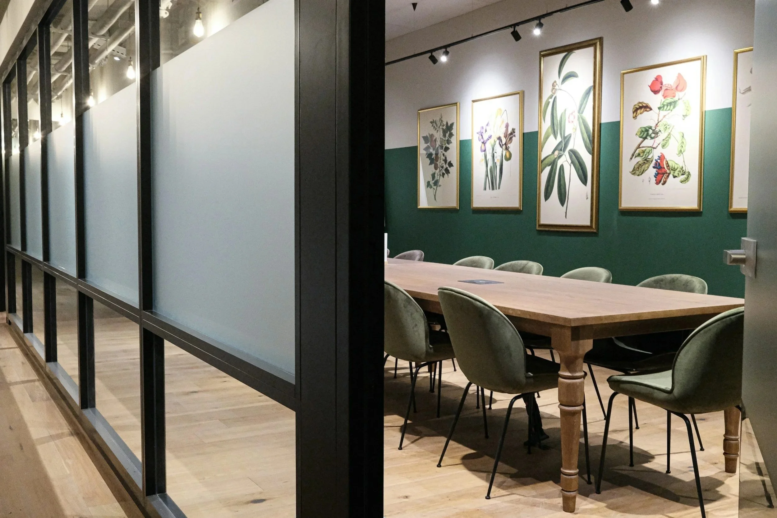 Interior of a modern art gallery or conference room featuring framed botanical artwork on a white and green wall, a large wooden table with green upholstered chairs, and decorative lighting. A glass partition with black framing is visible on the left side.