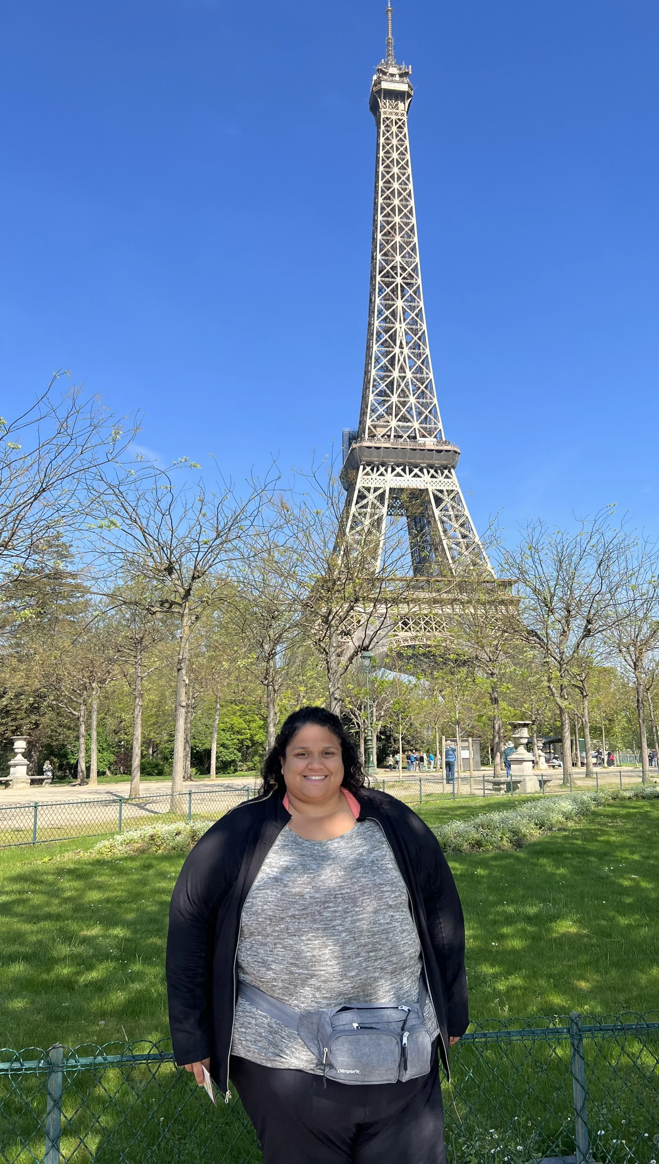 Woman standing in front of the Eiffel Tower in Paris, France, with trees and a park in the background on a sunny day.