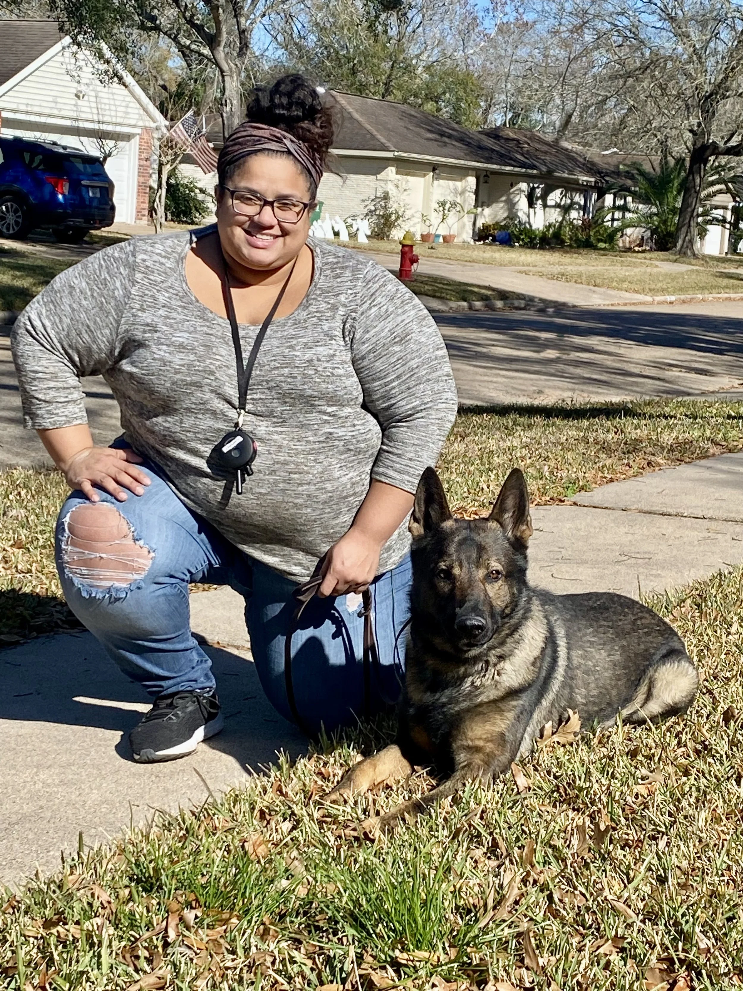 A woman with glasses, a headband, and ripped jeans squats on a sidewalk next to a German Shepherd dog with a black and tan coat. The woman is smiling and holding the dog's leash. The background shows a residential neighborhood with houses, a driveway, a blue car, trees with autumn leaves, a fire hydrant, and some potted plants.