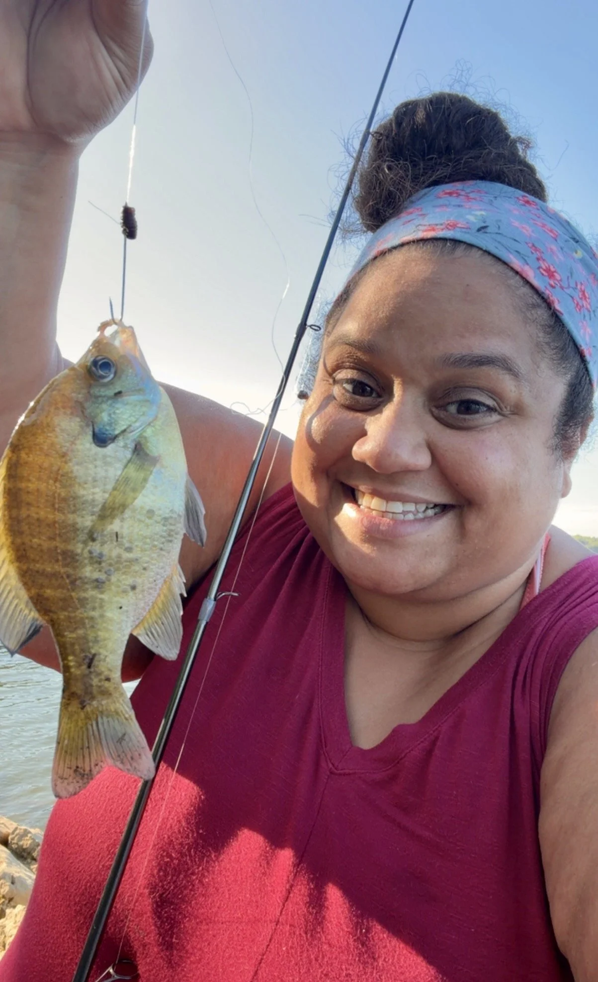 A woman smiling while holding a fish she caught, with a fishing rod in her hand, outdoors near a body of water, during the daytime.